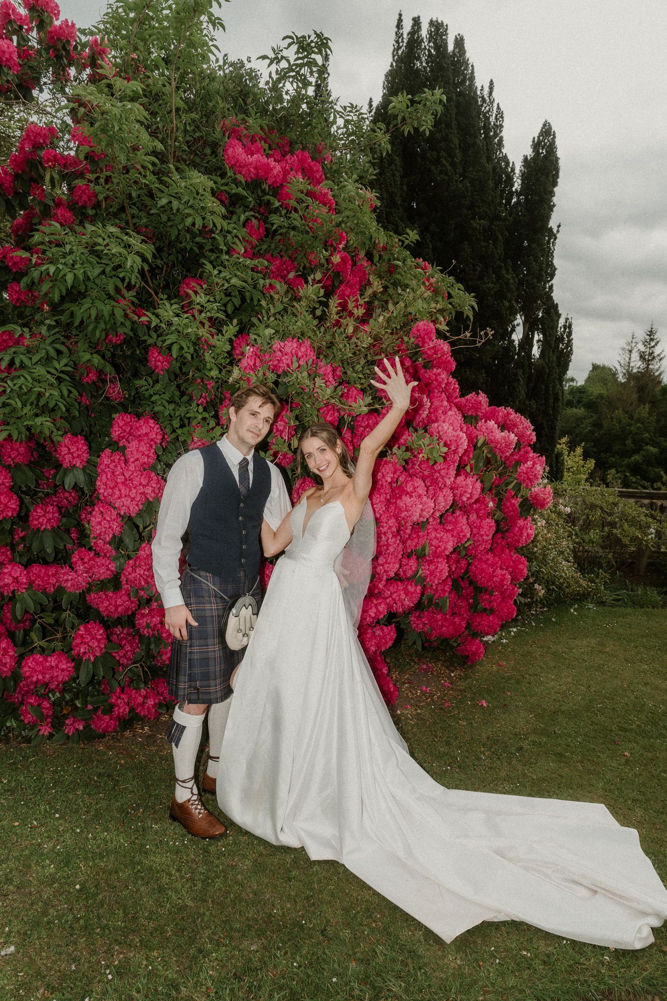 Bride and groom posing in front of large pink rhododendron bushes, groom wearing Scottish kilt, bride in white gown.