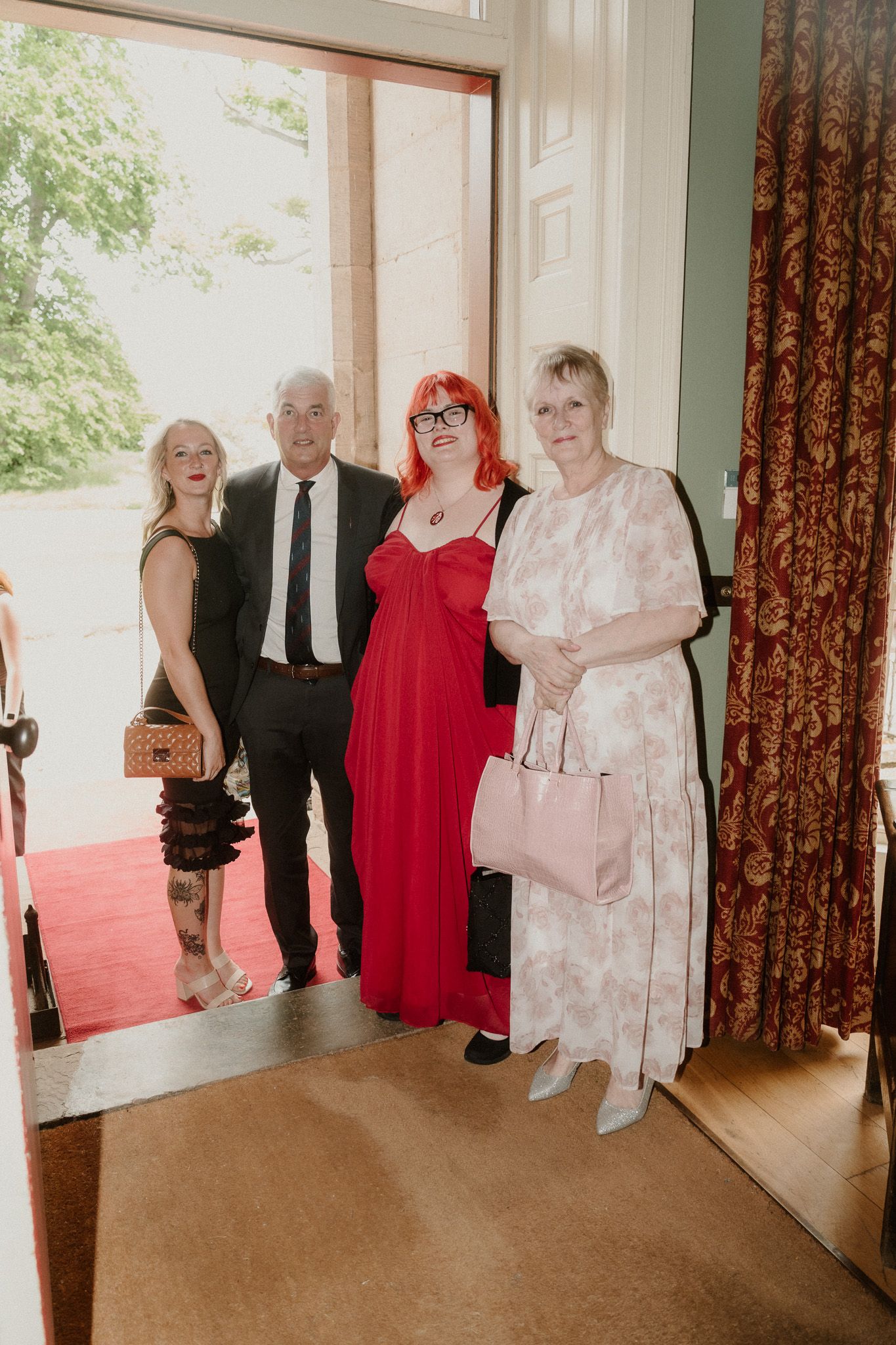 Four guests posing indoors at a formal event, one woman in a red gown with bright red hair, others in elegant attire.