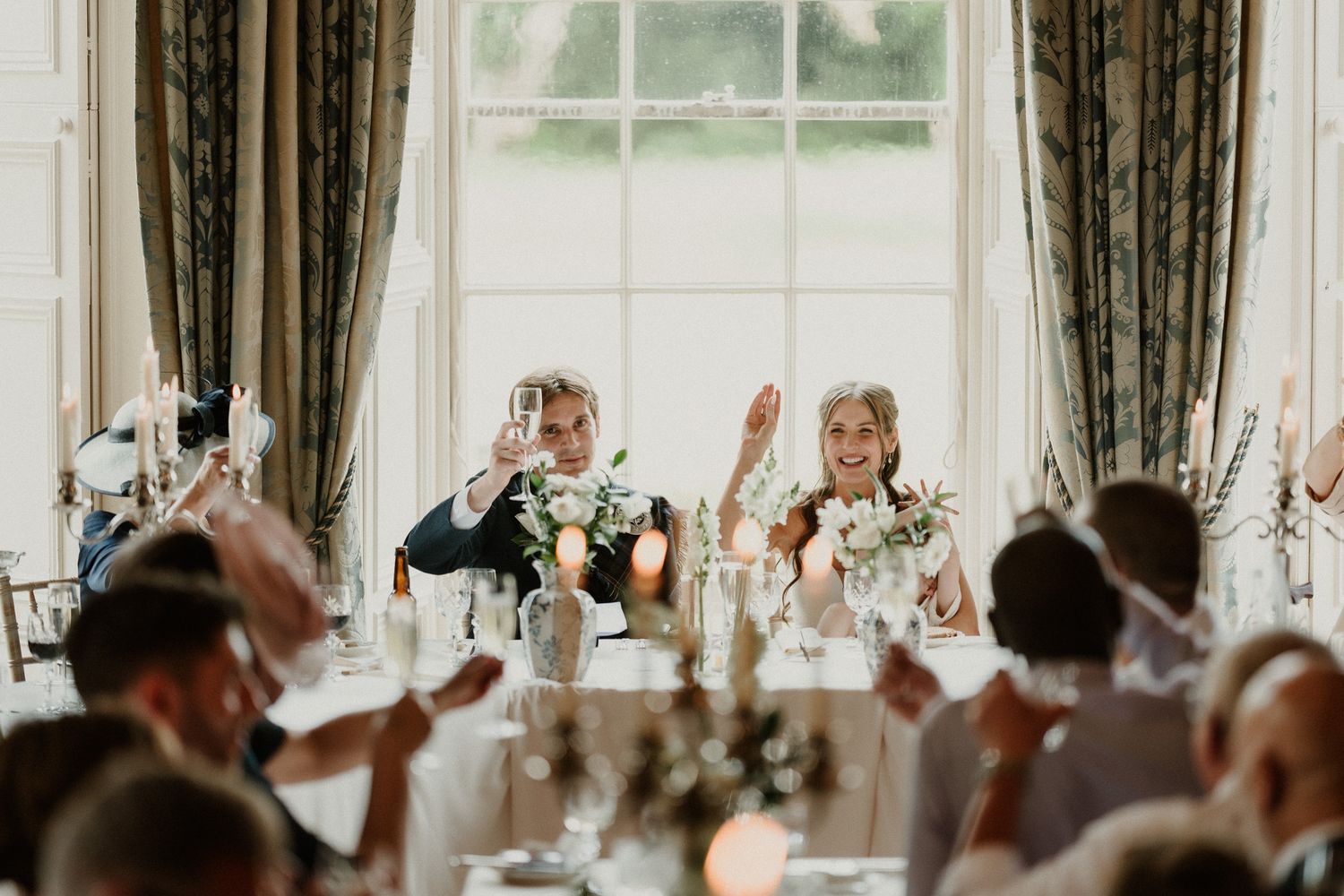 Bride and groom raise glasses during wedding reception toast at elegant venue with large windows.