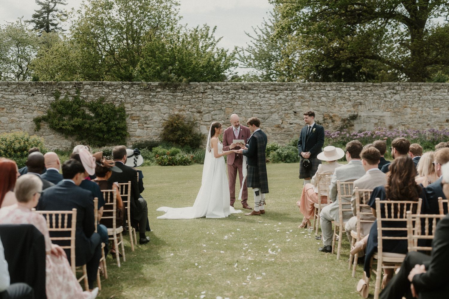 Outdoor wedding ceremony in a walled garden with guests seated on wooden chairs as couple exchanges vows.