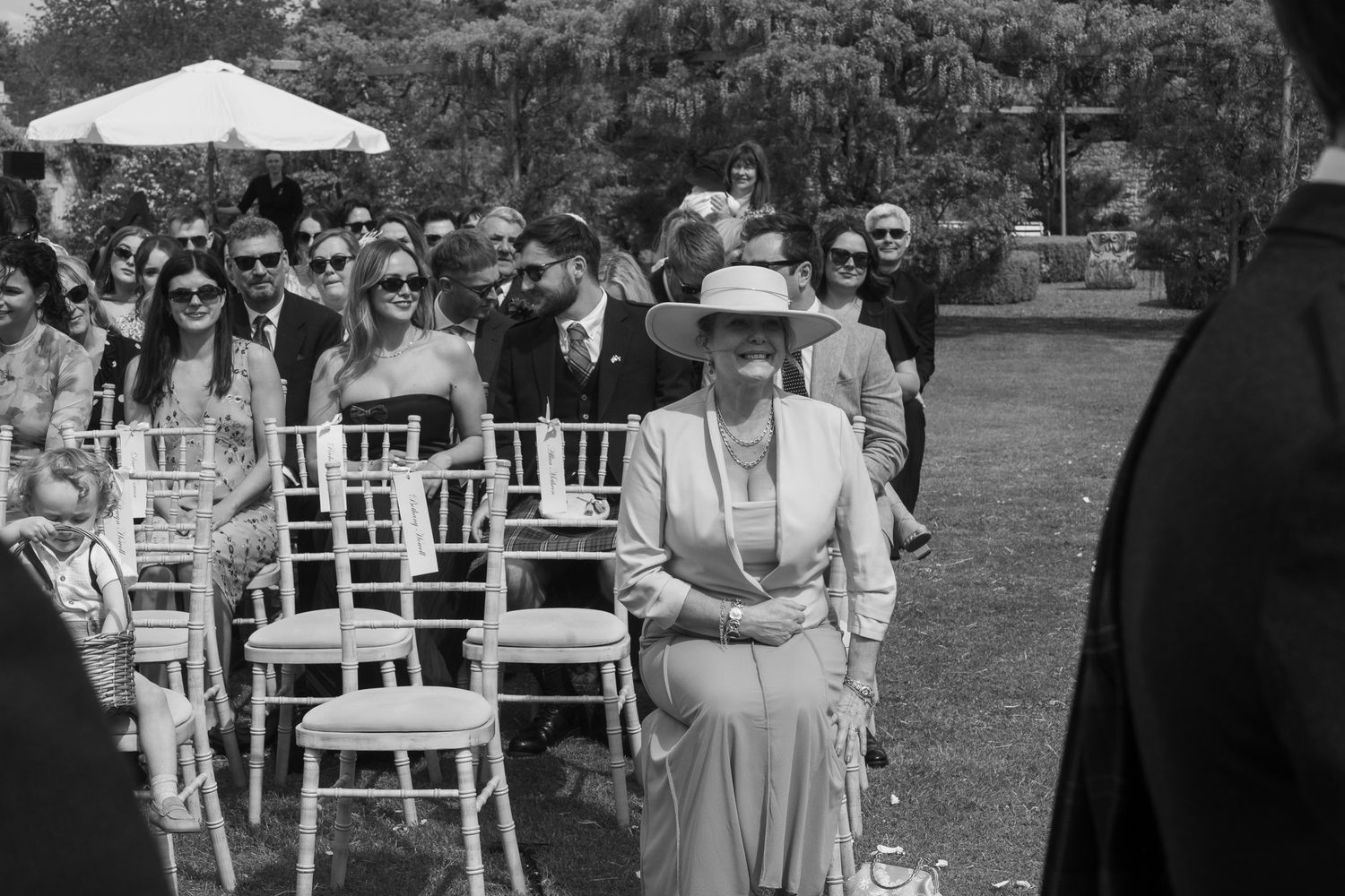 Black and white outdoor wedding ceremony with guests seated on white chairs, an elegant woman in hat standing in aisle.