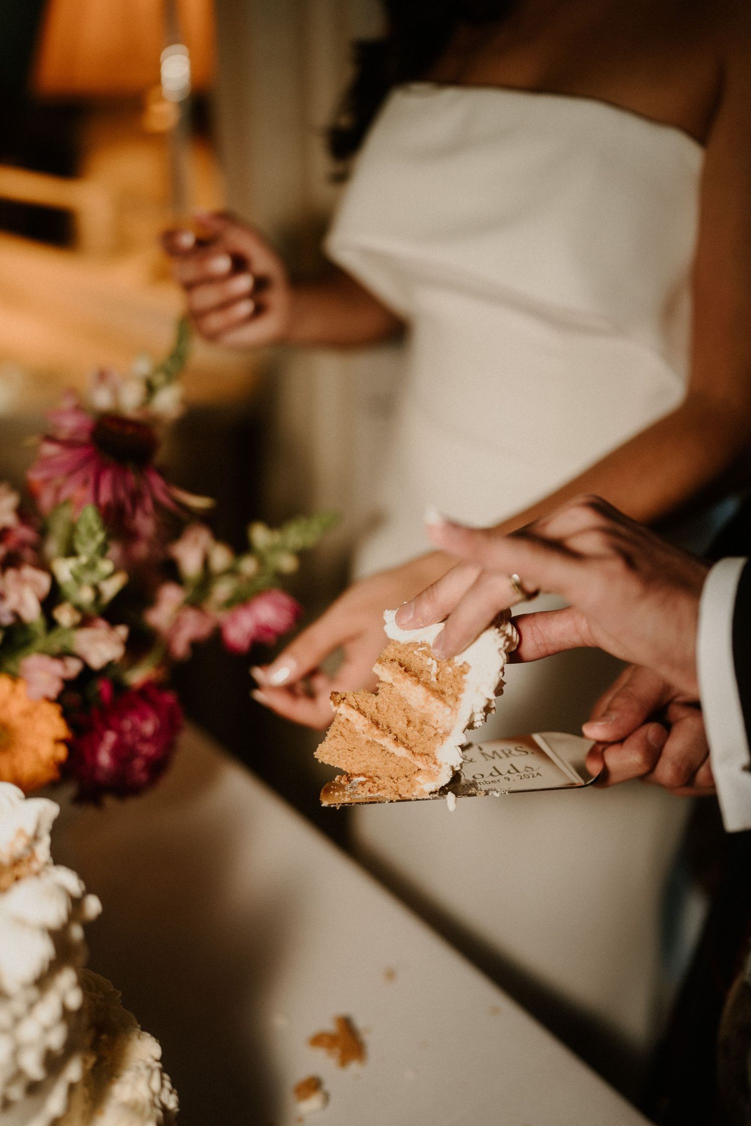 Bride and groom cutting a layered wedding cake with white frosting, surrounded by colorful floral arrangements.