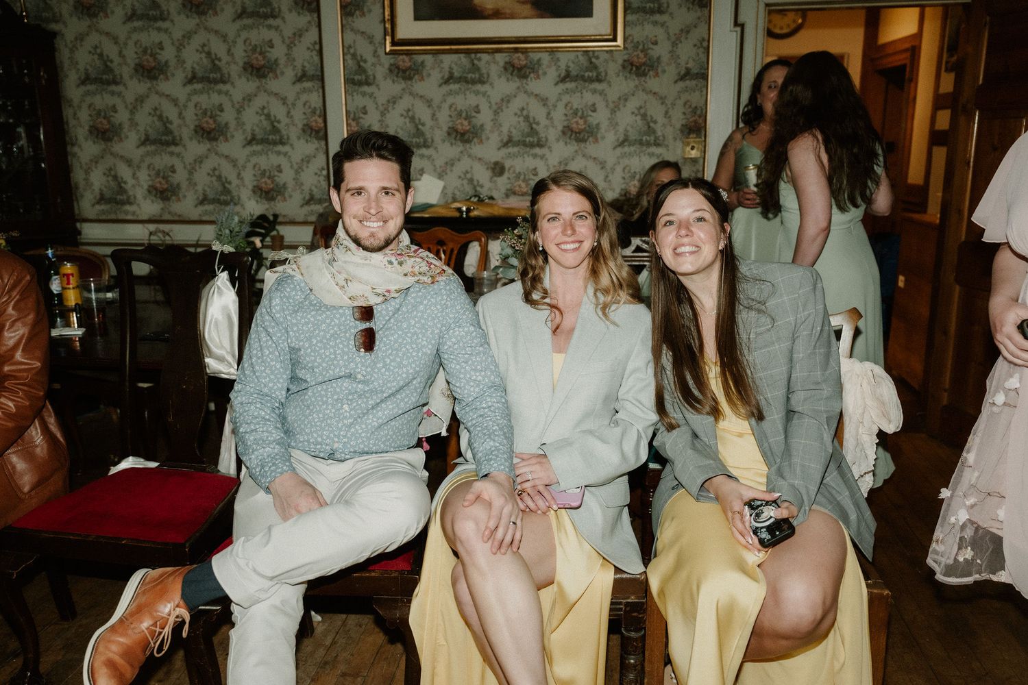 Three friends smiling and posing together at an elegant indoor social gathering with floral wallpaper backdrop.