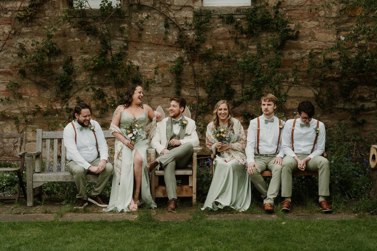Wedding party laughing on a bench in front of a stone wall, bridesmaids in sage green dresses and groomsmen in suspenders.