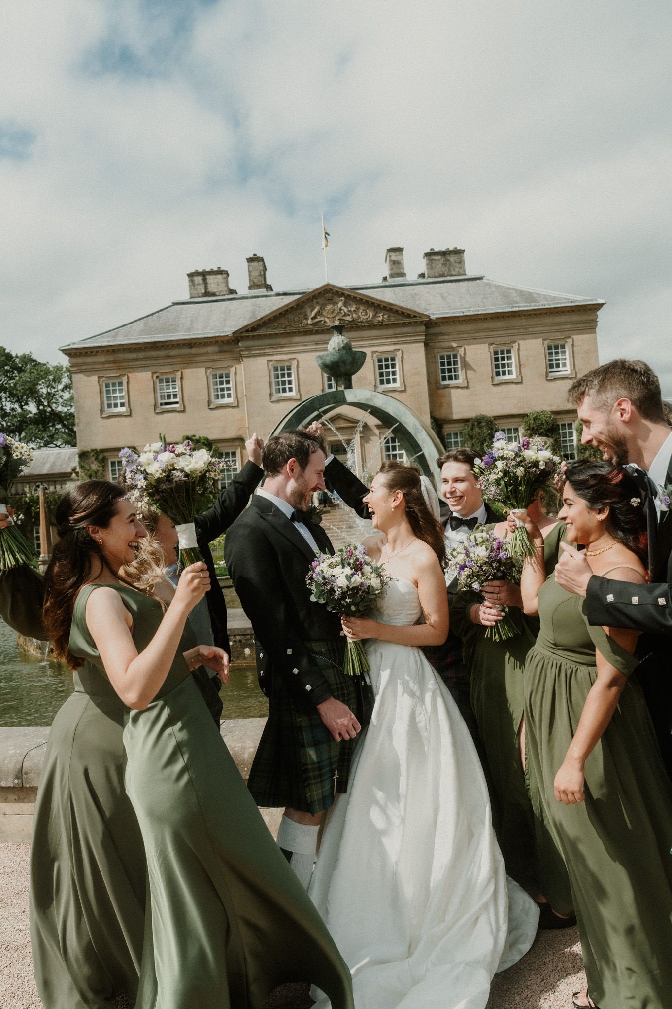 Bride and groom share a moment surrounded by bridesmaids in sage green gowns outside an elegant Scottish manor.