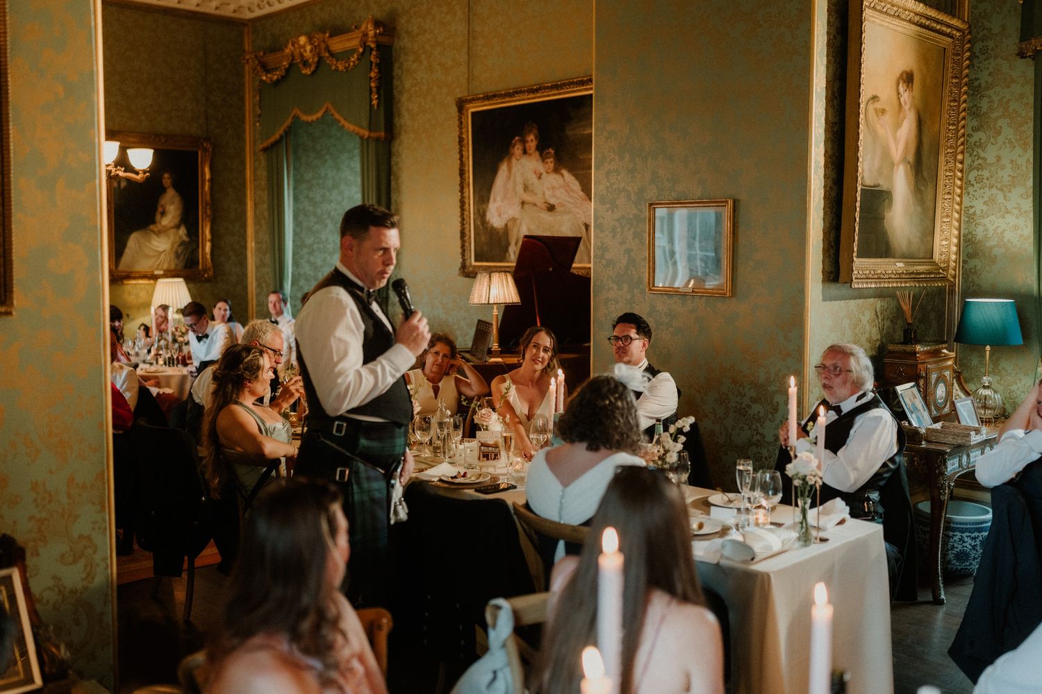 Man in vest gives a speech holding a microphone as wedding guests dine at candlelit tables in an elegant room with ornate framed portraits.