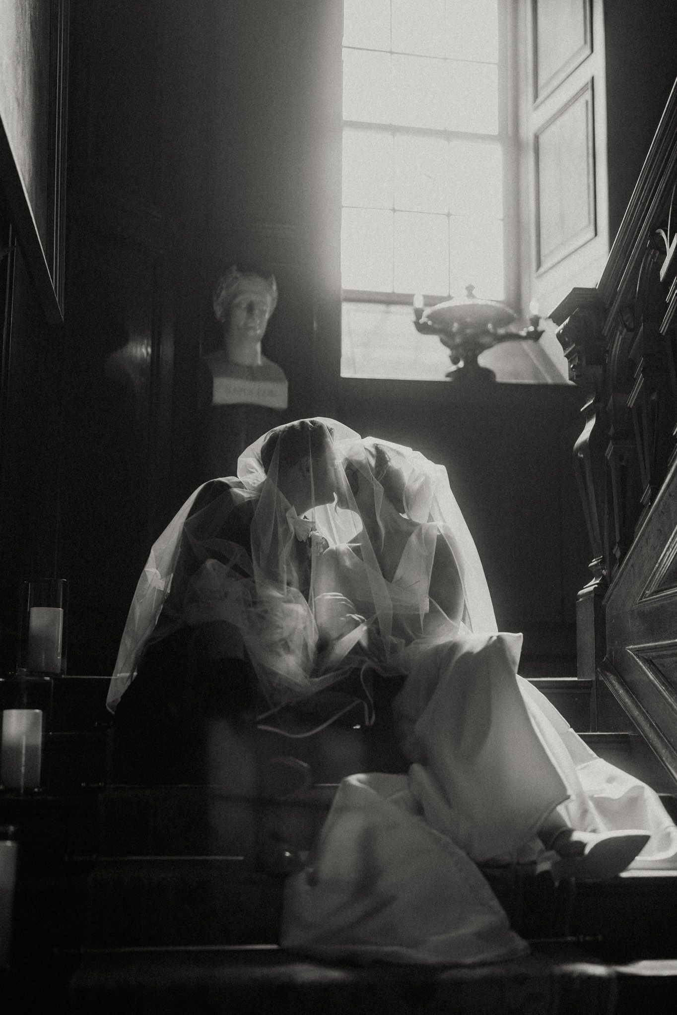 Black and white photo of a couple kissing beneath a sheer veil on a grand staircase, with a classical bust and window light behind.