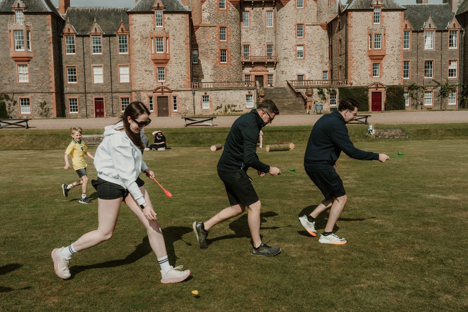 Group of people playing a lawn game with sticks on a green lawn in front of a historic stone castle with turrets on a sunny day.