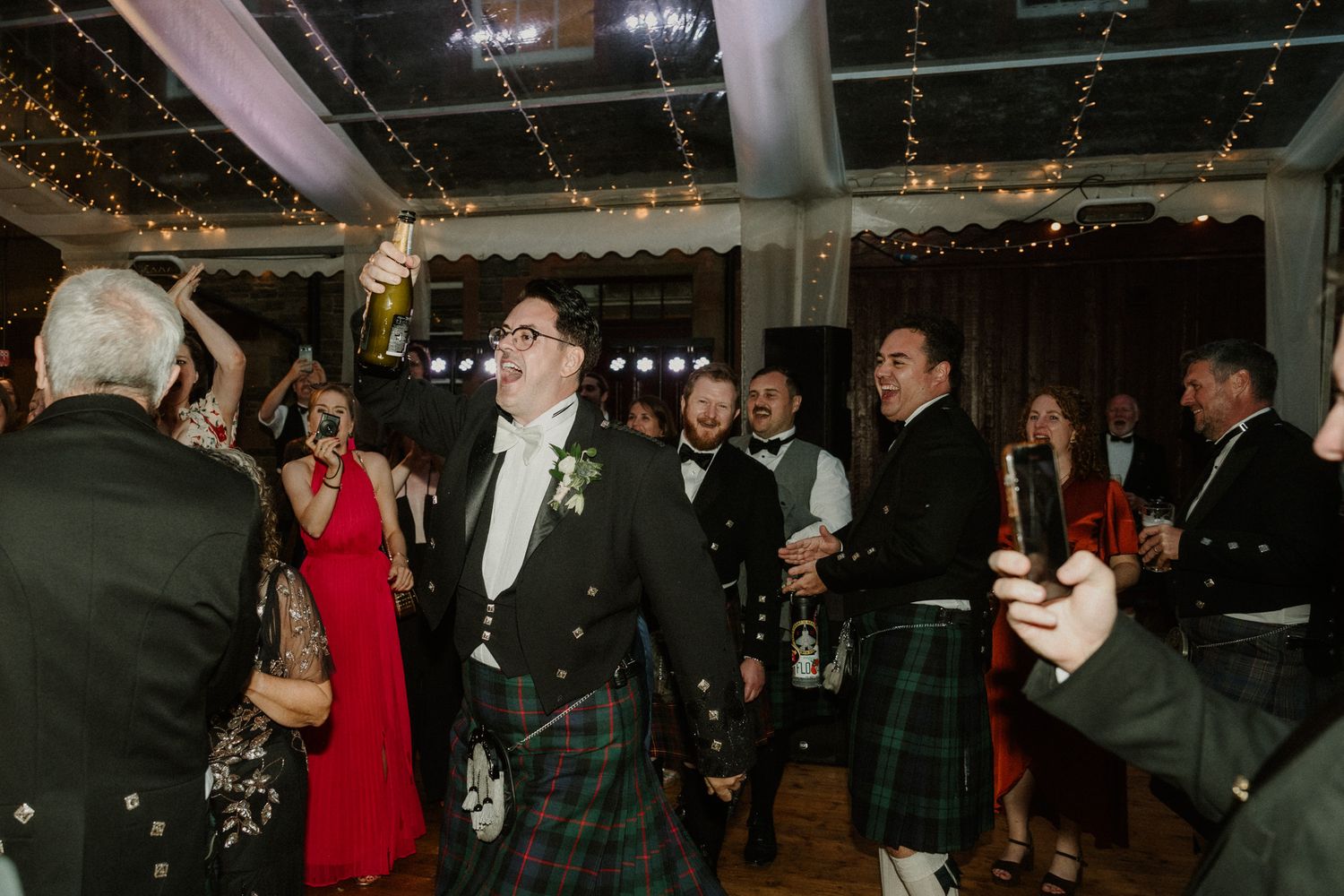 Guests in formal kilts and evening wear dance and celebrate at a lively Scottish wedding reception under string lights.