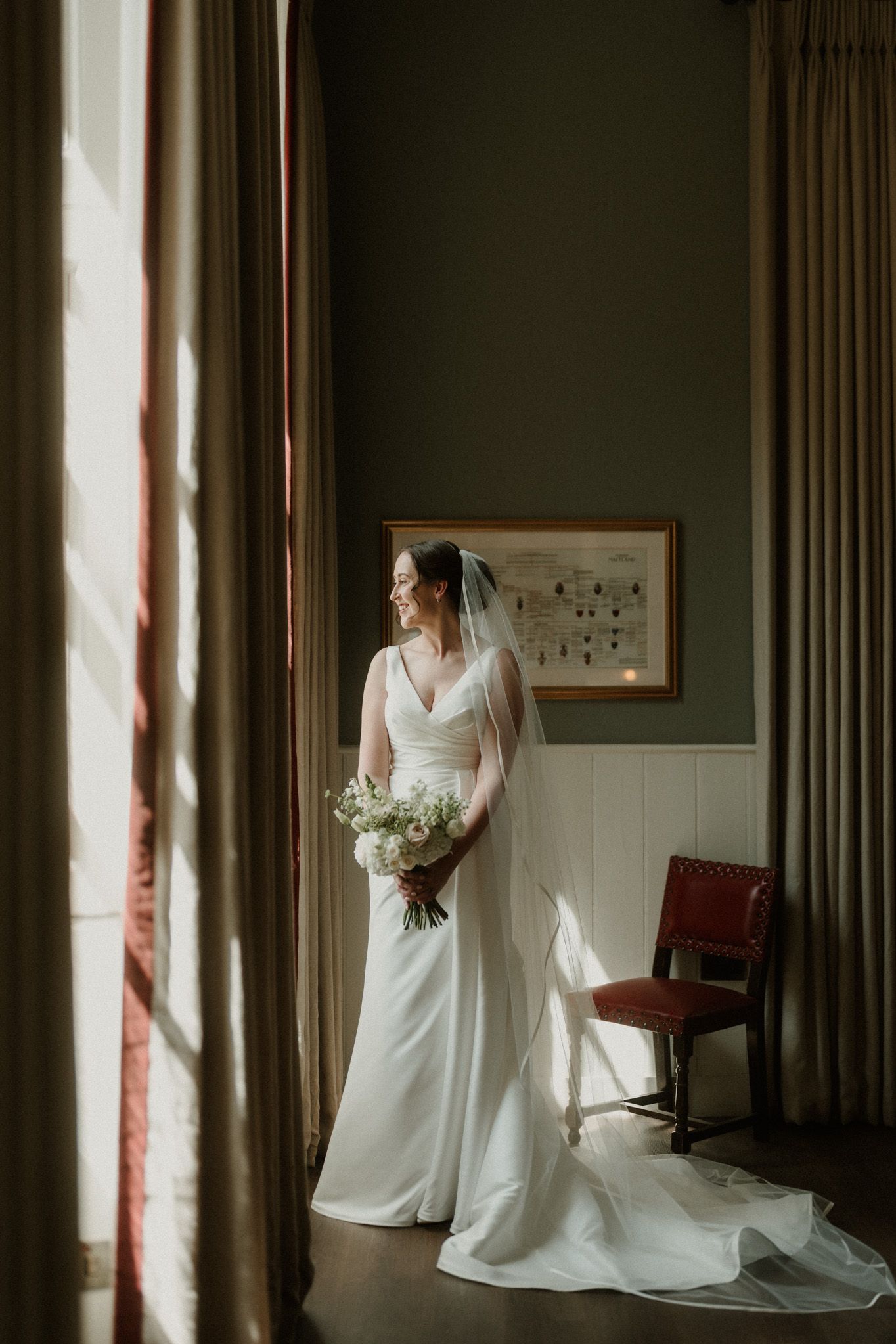 Bride in an elegant white gown and long veil holds a white bouquet beside a sunlit window with drapes in a classic room with a red chair.