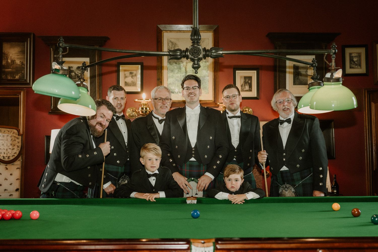 Six men and two boys in black tuxedos and bow ties pose around a green snooker table in a vintage room with red walls and green lamps.