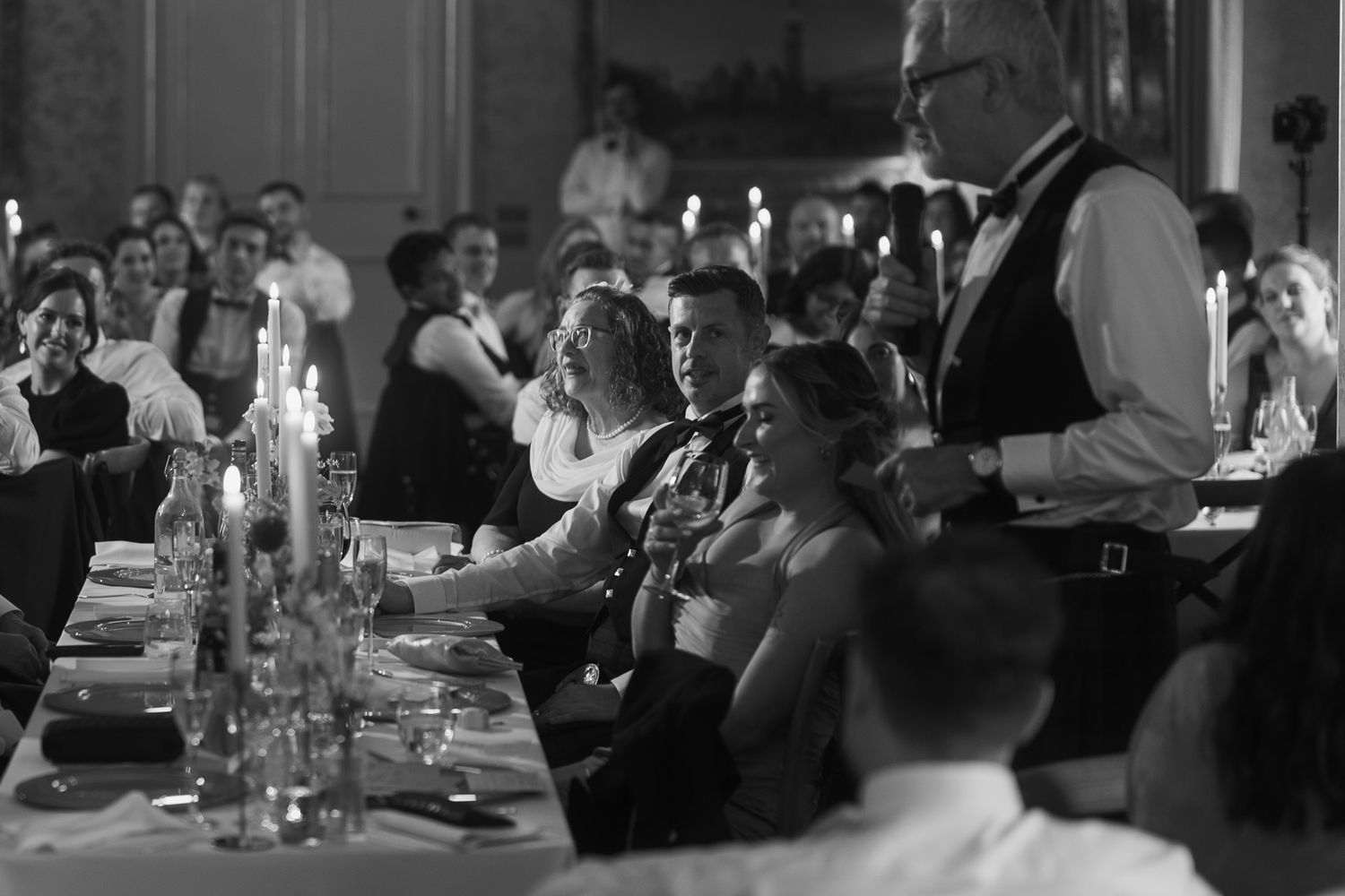 Guests seated at a candlelit banquet table laugh and listen as a man in a vest stands to give a toast in a black and white photo.