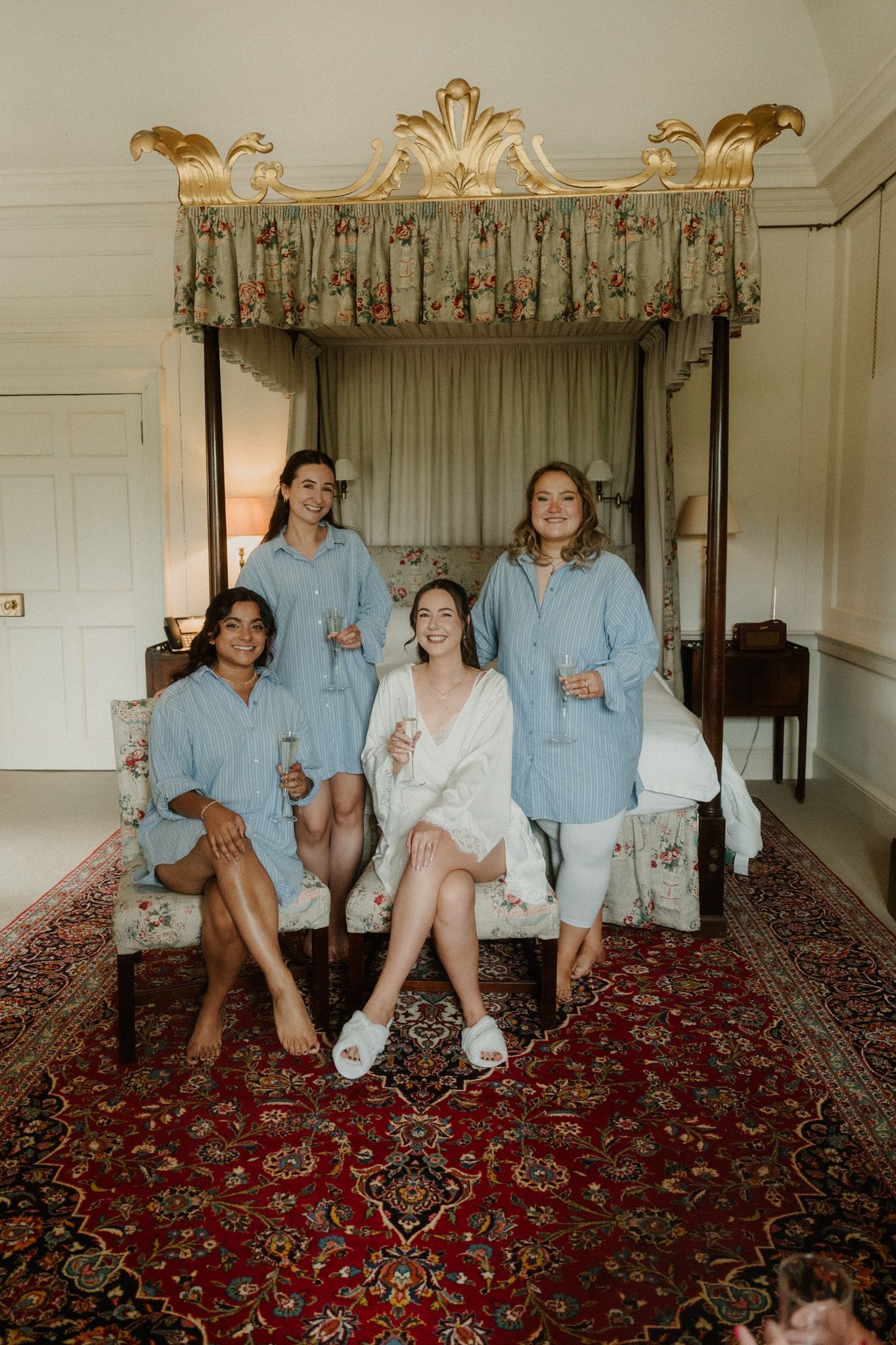 Bride in white robe with three bridesmaids in blue robes celebrating with champagne in ornate bedroom.