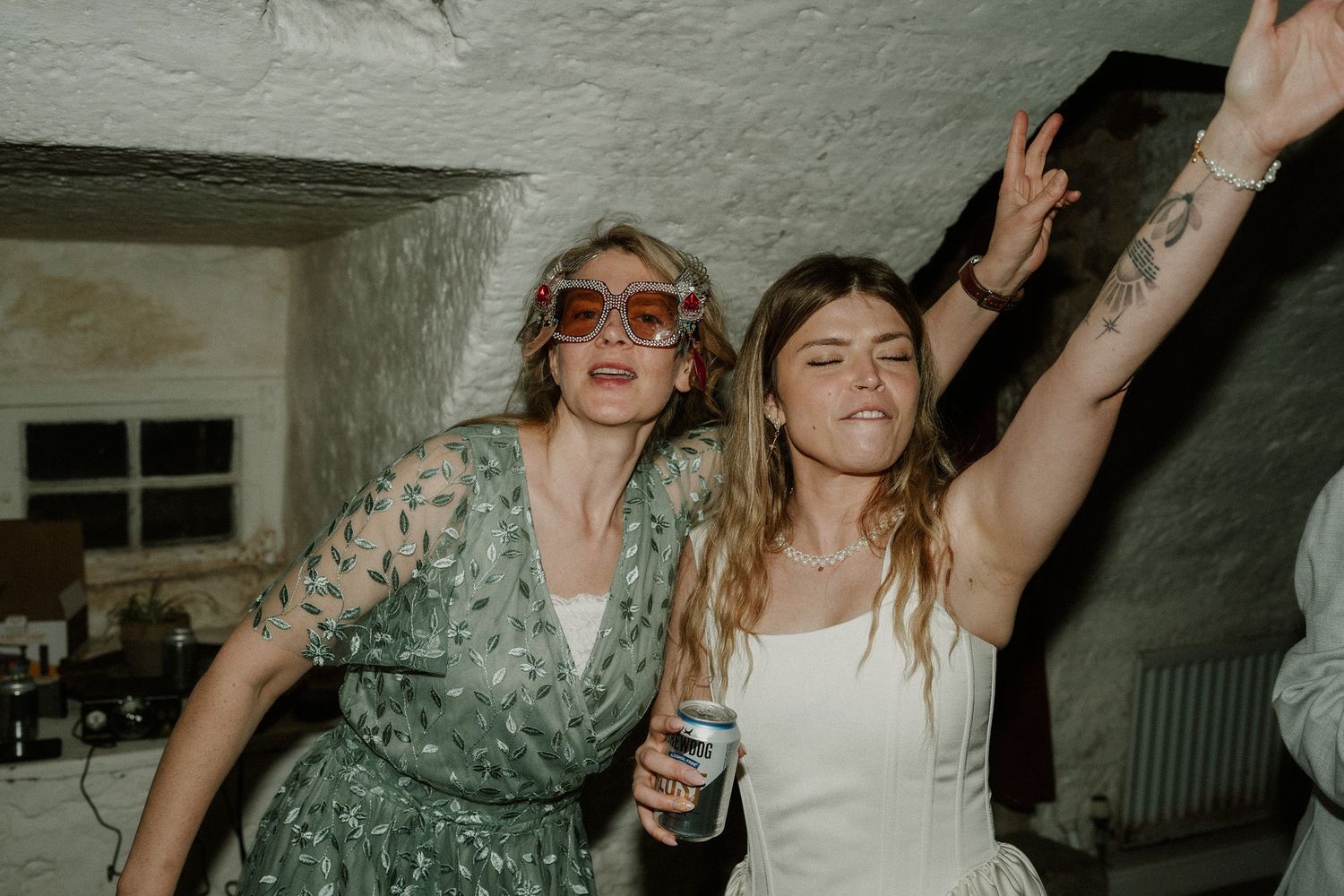 Two women dancing joyfully at a party, one in a floral dress with pink sunglasses, the other in white.