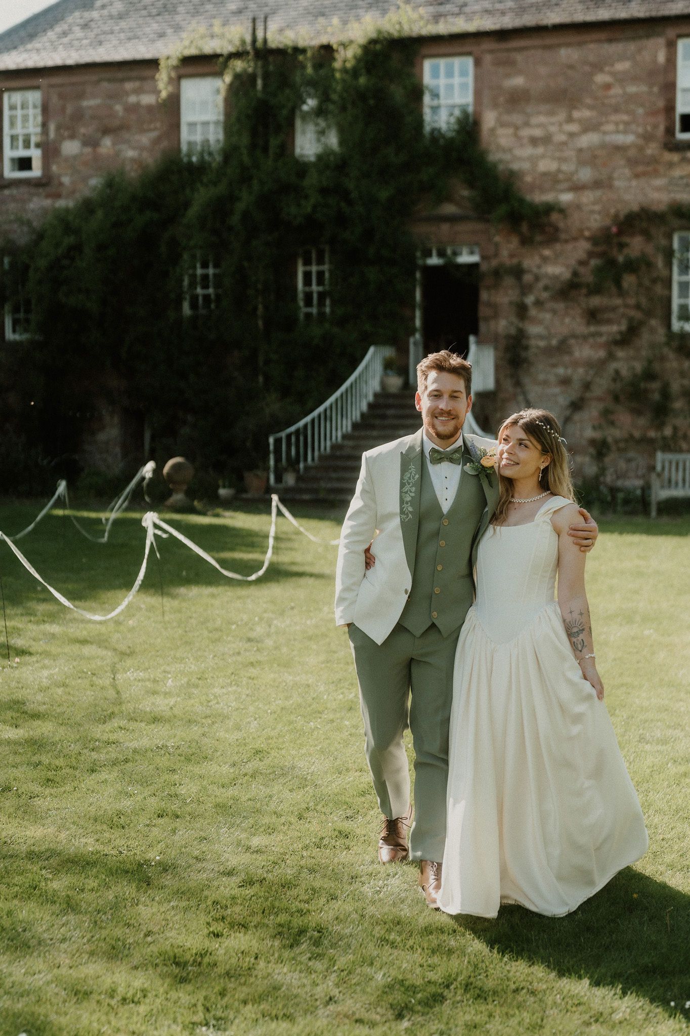 Bride and groom walking on green lawn outside ivy-covered stone manor house on sunny wedding day.