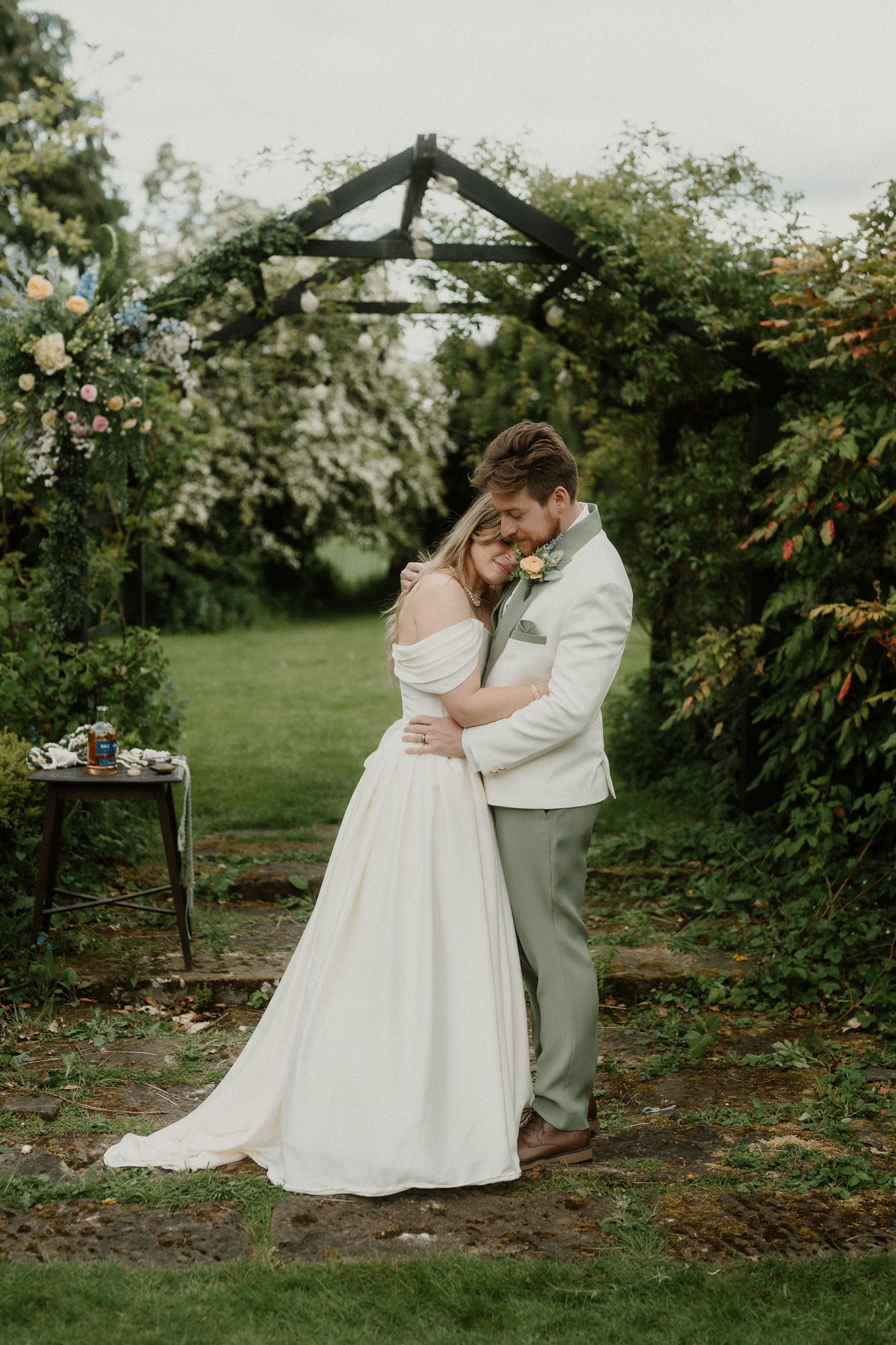 Bride and groom embrace under a floral garden arch in a lush outdoor wedding ceremony setting.