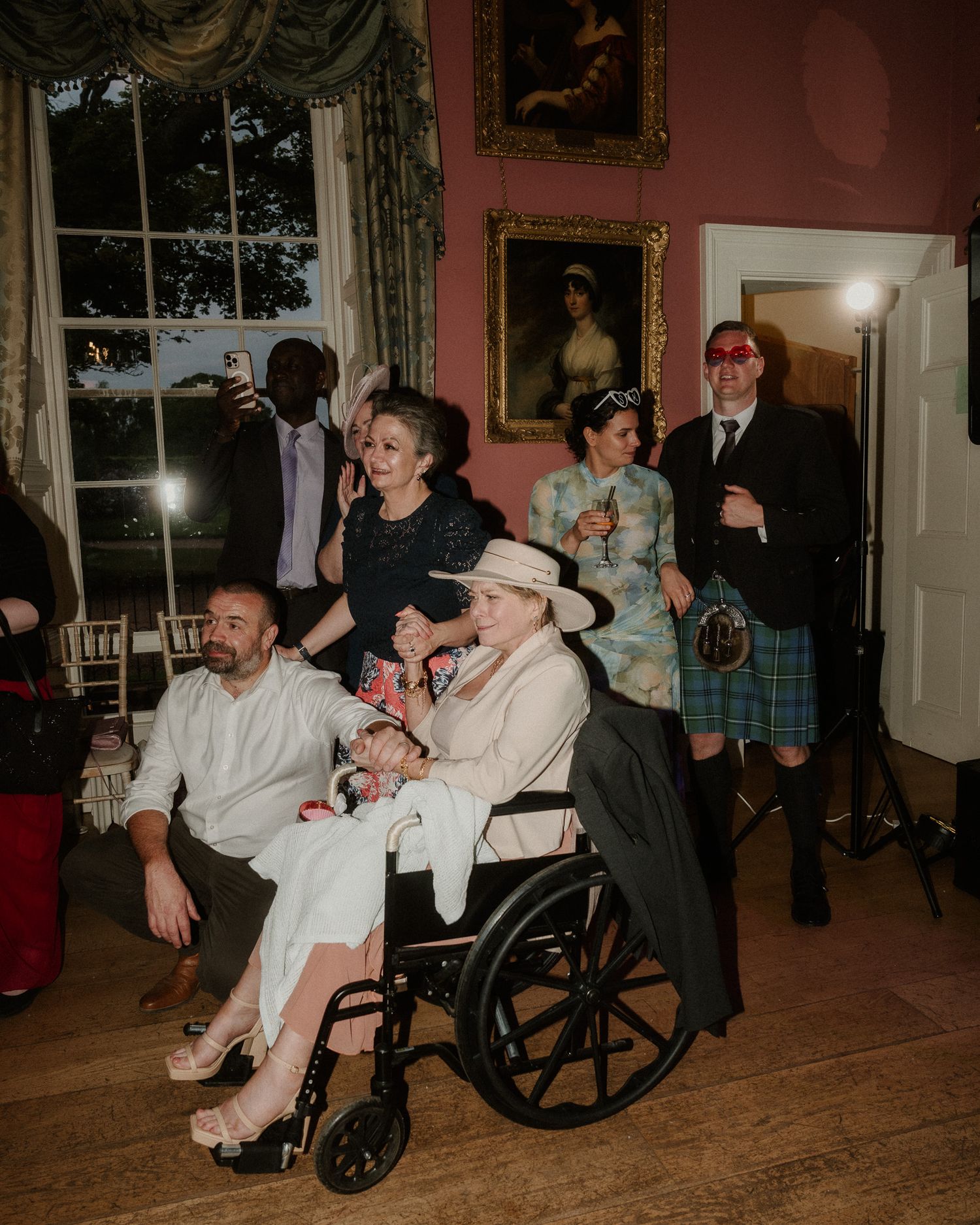 Elegant gathering in a pink room with guests in formal attire, including a woman in a wheelchair, at a special event.