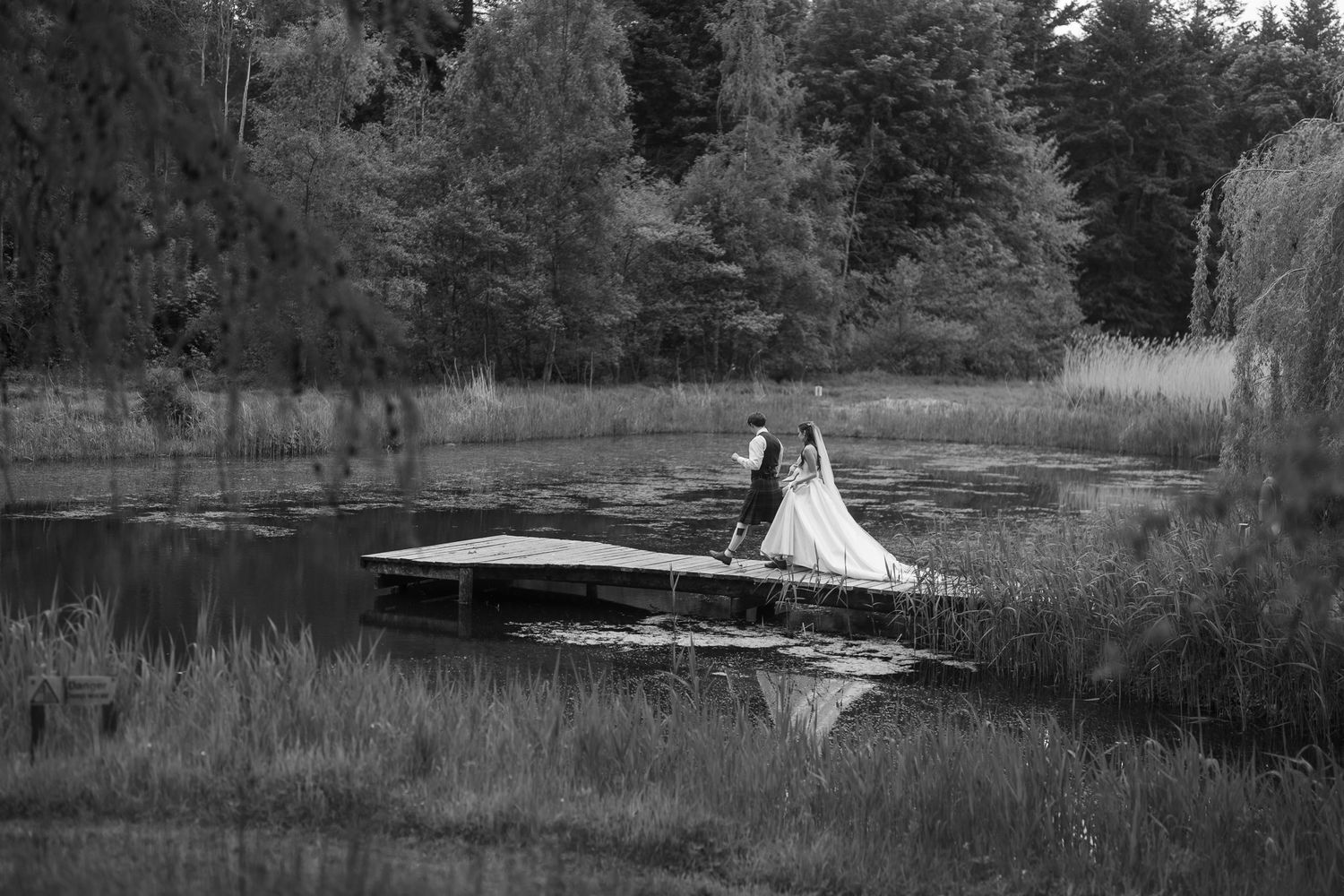 Black and white photo of a bride and groom standing on a dock surrounded by a serene pond and lush trees.
