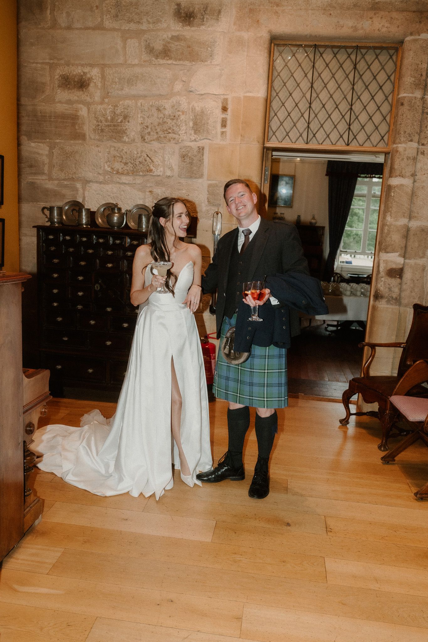 Bride in white gown and groom in blue tartan kilt holding wine glasses in a stone-walled Scottish castle venue.