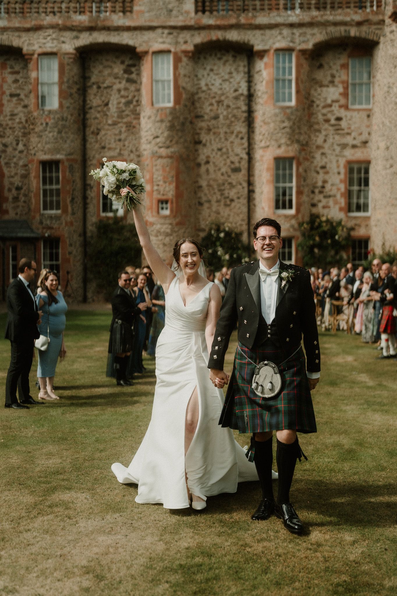 Smiling bride in a white gown raises her bouquet beside a groom in a tartan kilt as wedding guests cheer on a lawn before a stone building.