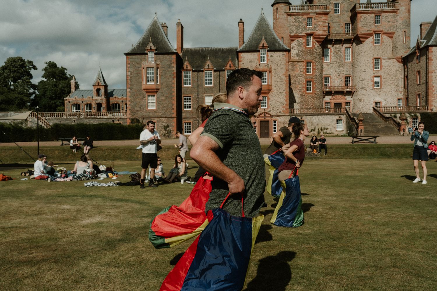 A man in a green t-shirt takes part in a sack race on a sunny lawn in front of a historic stone castle while spectators look on.