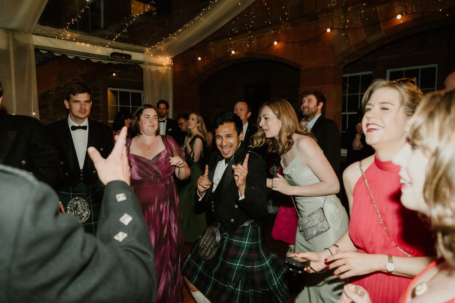 Wedding guests in formal attire and tartan kilts clap and dance together at a lively indoor reception with warm string lighting.