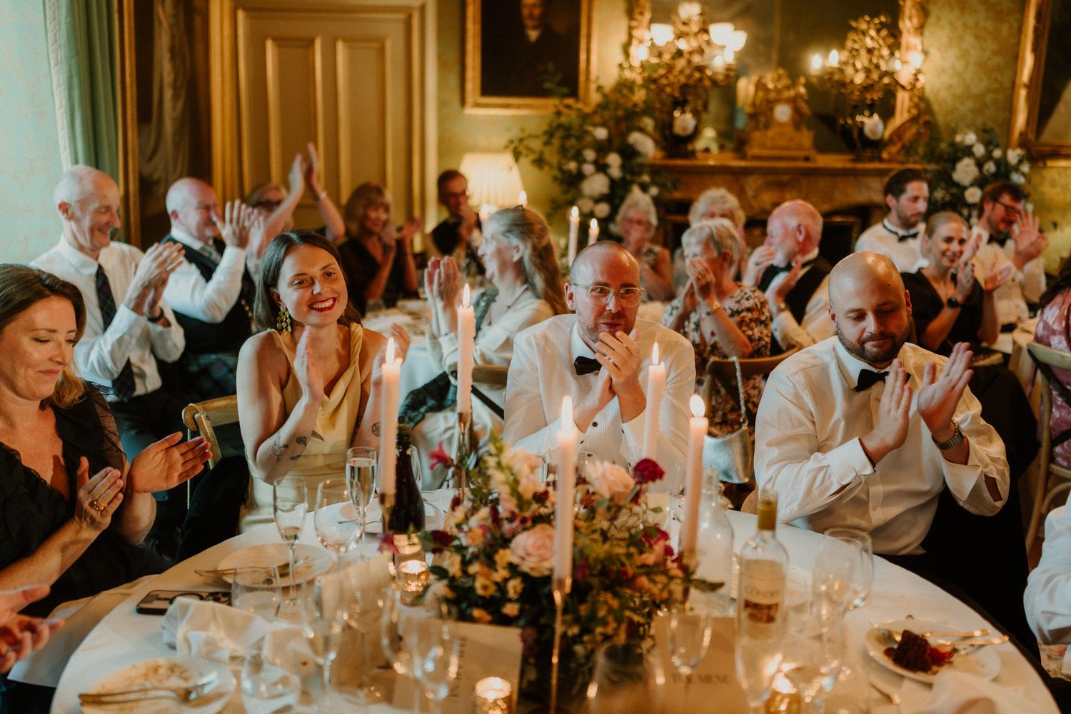Wedding guests laugh and applaud at a candlelit banquet table with flowers, glassware, and an ornate vintage room in the background.