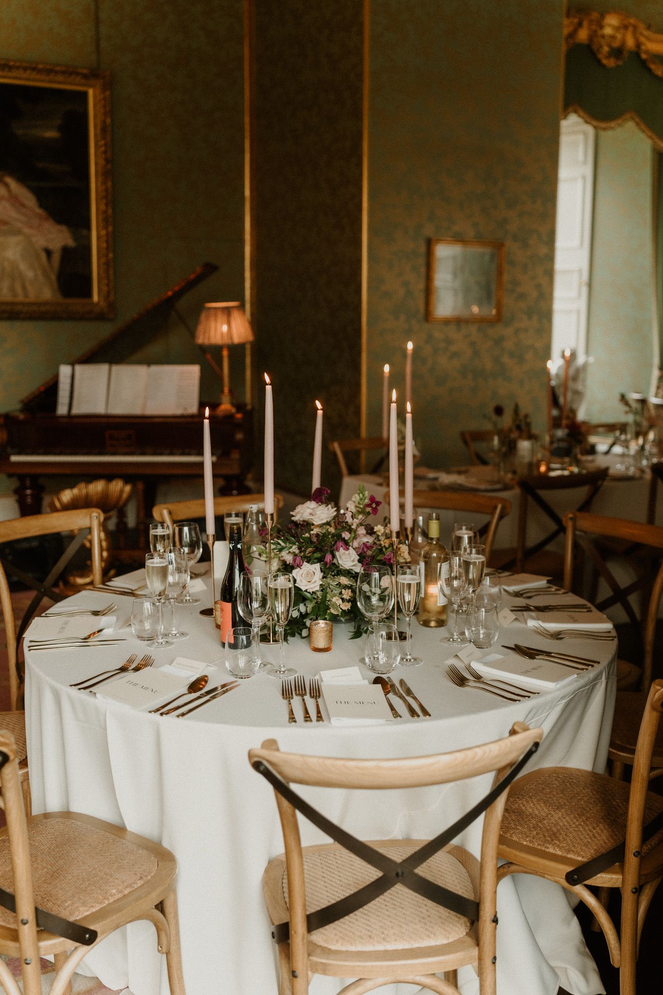 Elegant wedding reception table with white linens, tall taper candles, floral centerpiece and cross-back chairs in a historic green room.