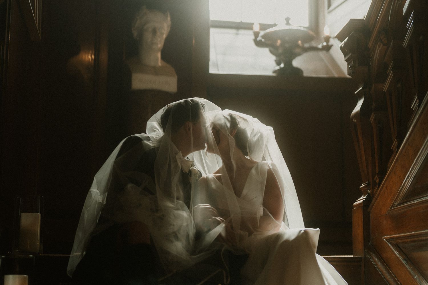 Two brides in white wedding gowns and veils share an intimate moment seated together in a warmly lit vintage room.