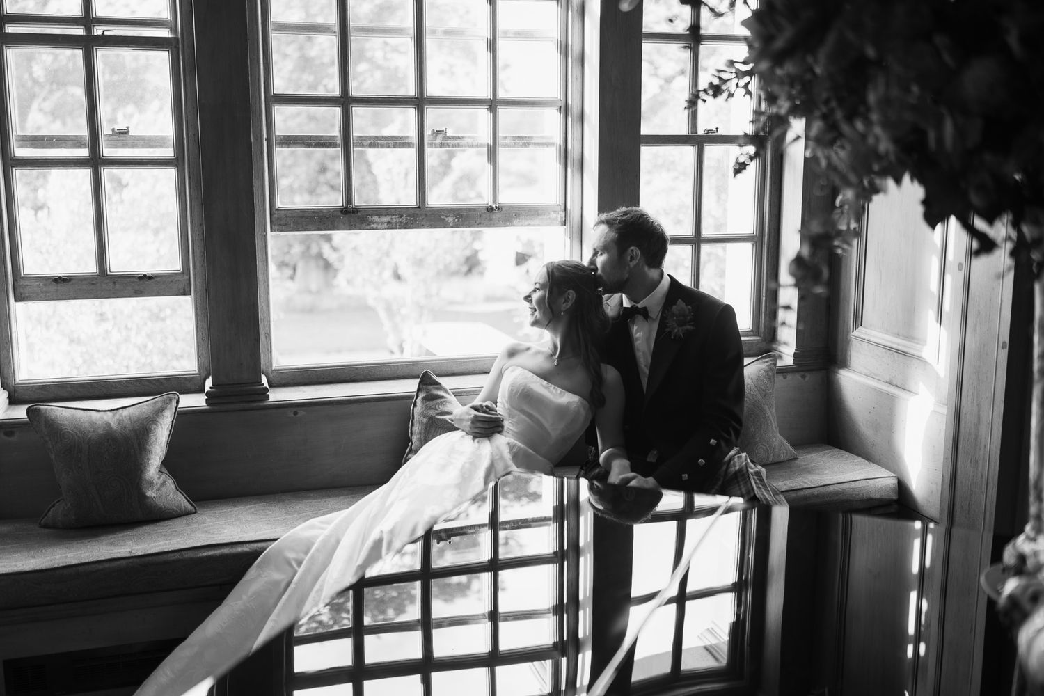 Black and white photo of a bride and groom sharing a romantic moment by large windows in an elegant venue.