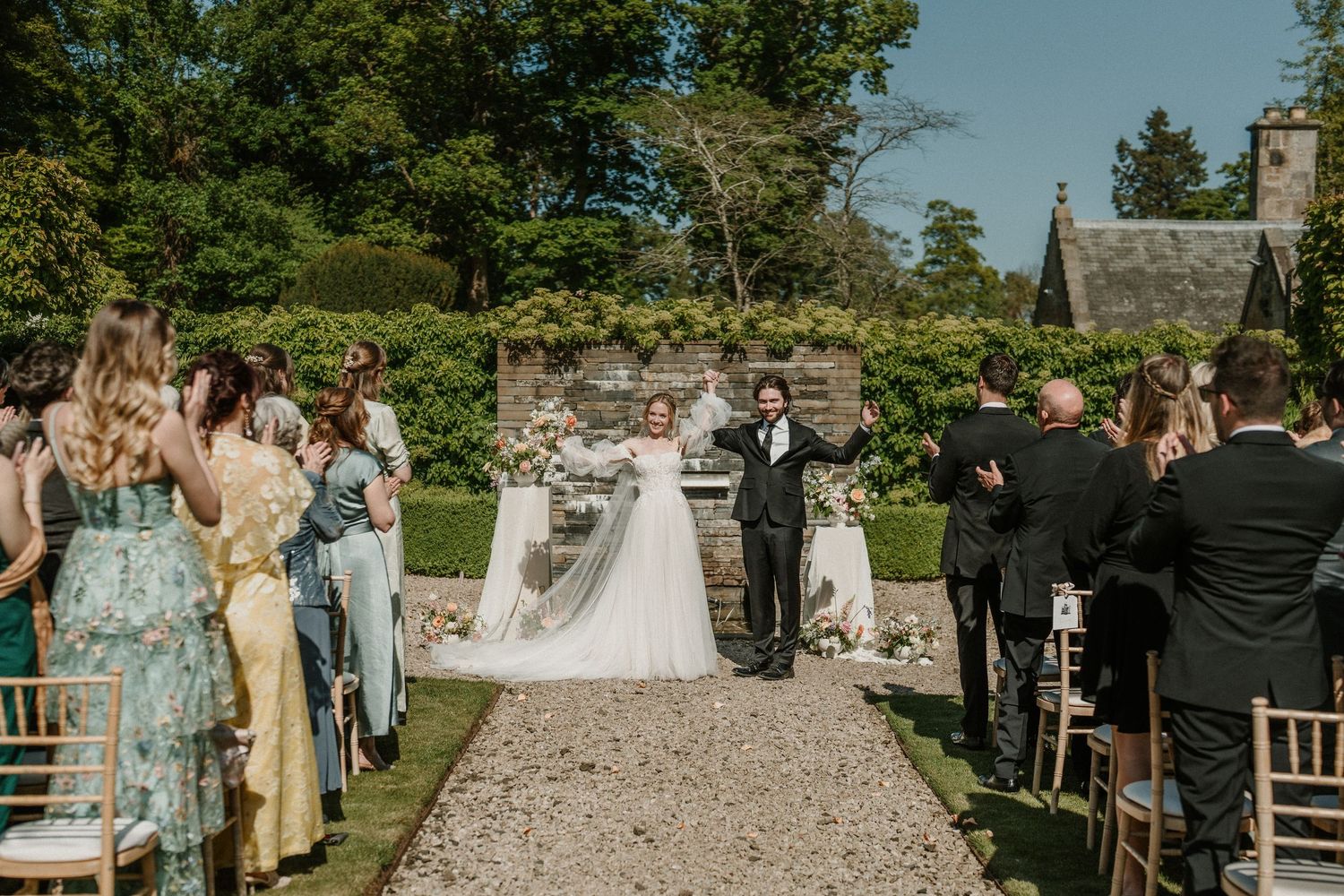Elegant outdoor wedding ceremony with guests seated on white chairs along a stone pathway.