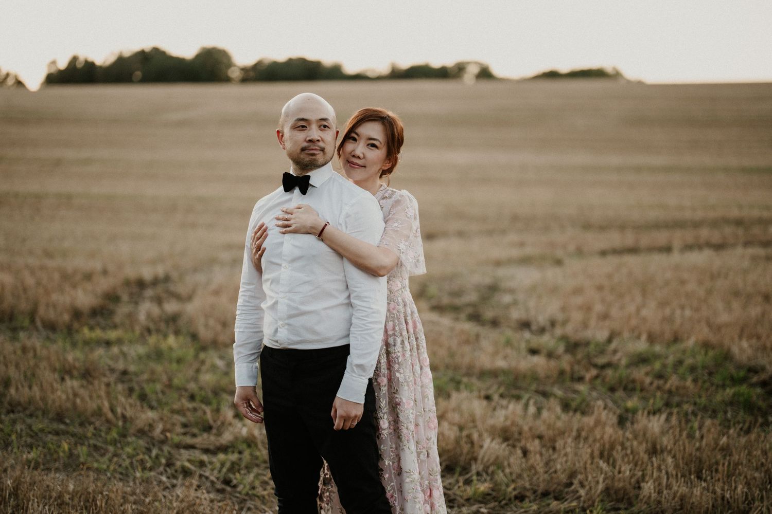A couple embraces while standing in a vast wheat field at golden hour.