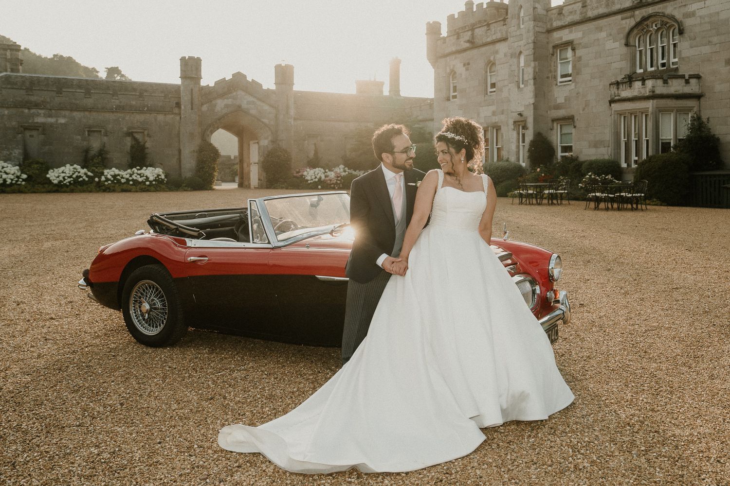 Elegant wedding photoshoot with a vintage red convertible car against the backdrop of a castle at golden hour.