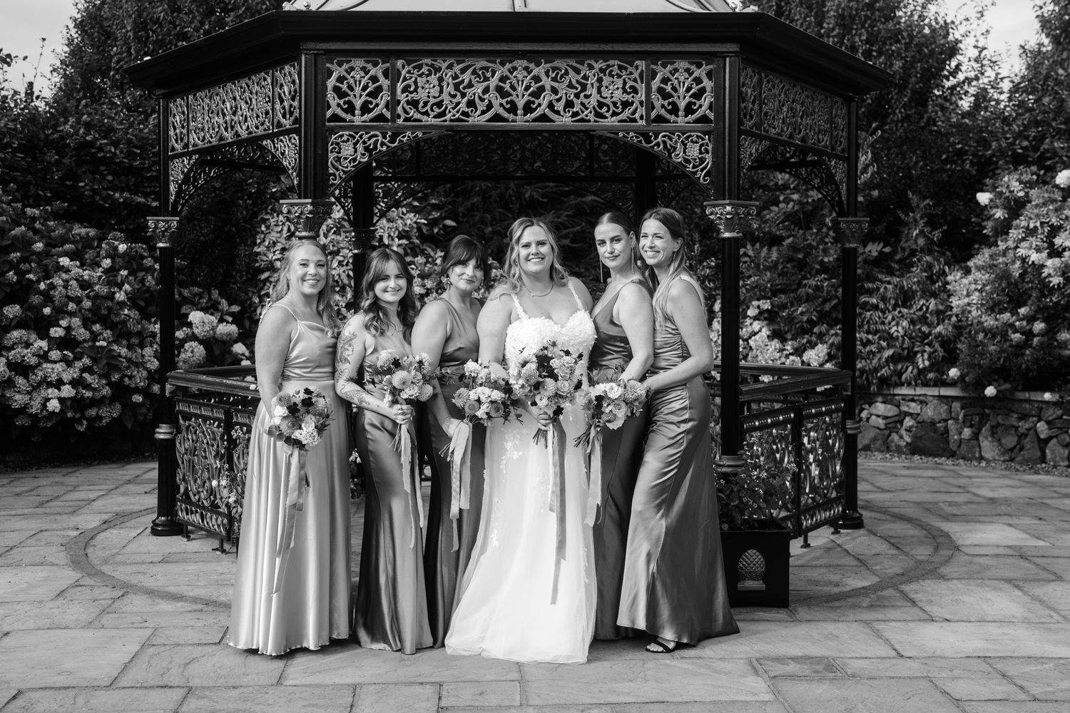 Wedding party lined up for photos under an ornate garden gazebo.