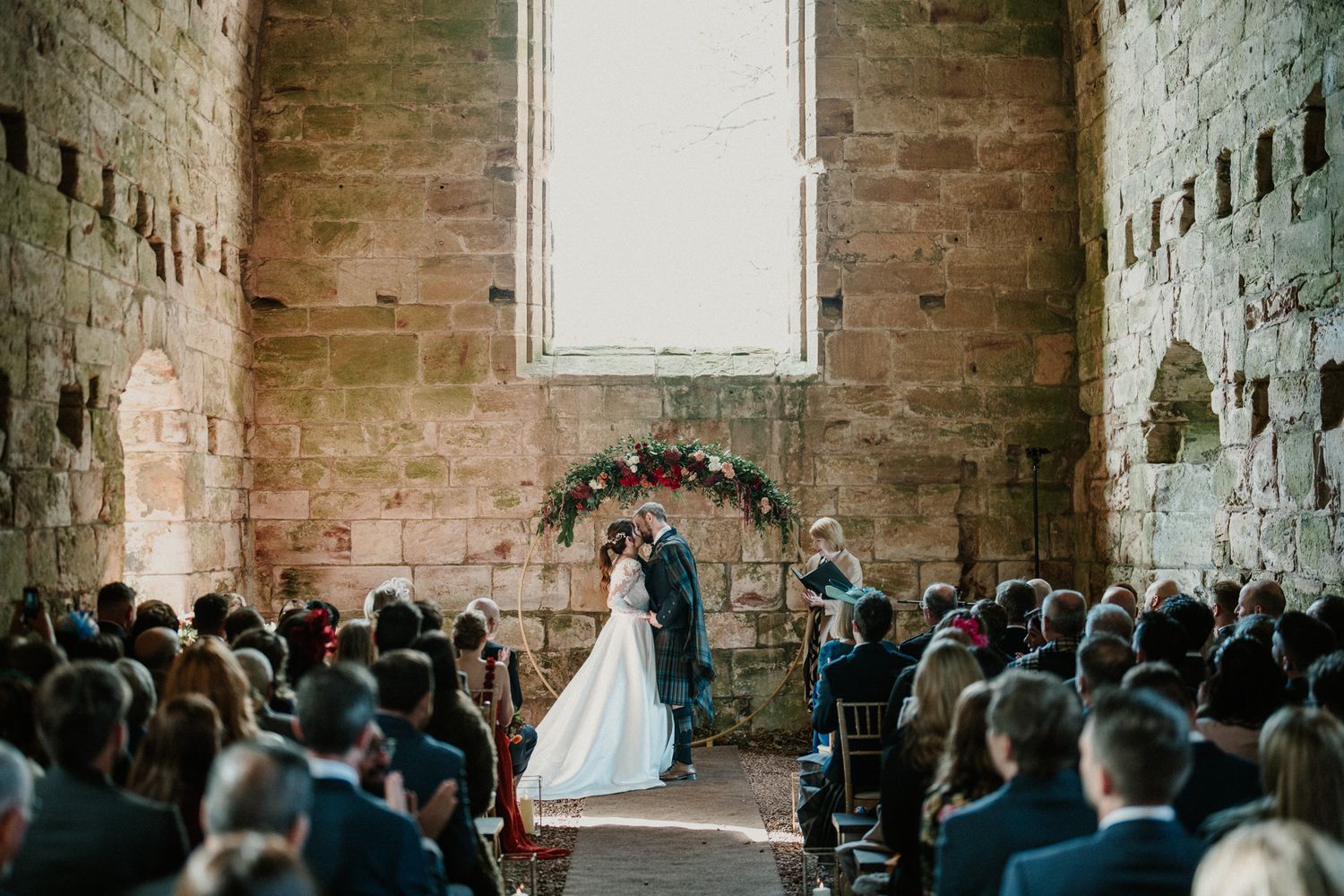 Wedding ceremony in a stone-walled chapel with guests seated in rows beneath a tall arched window.