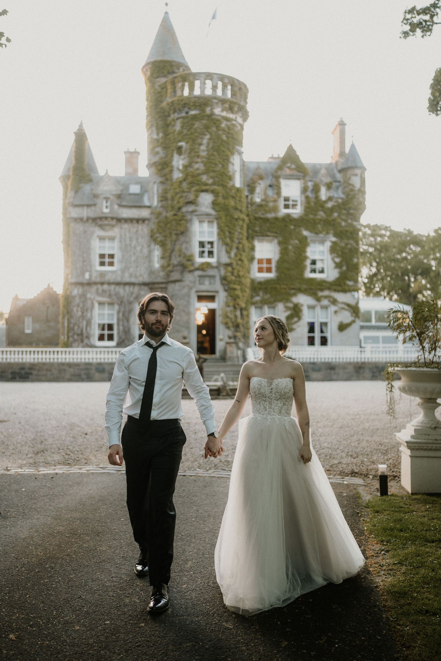 Newlyweds stroll together on the grounds of an ivy-covered castle at dusk.