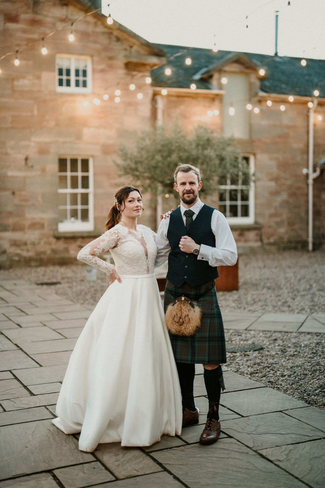 Newlyweds sharing a playful moment on stone patio with string lights above.