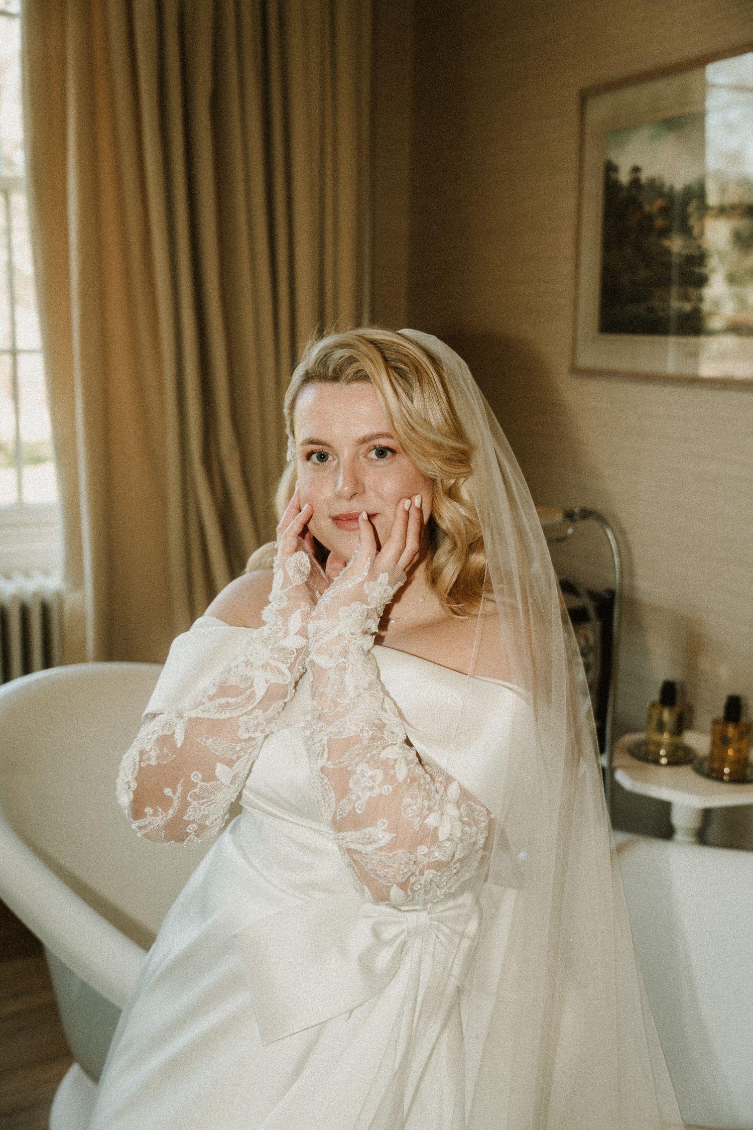 A bride in a white lace gown and veil poses emotionally in a sunlit room.