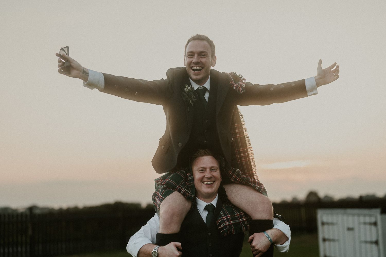 Two people in Scottish formal wear strike playful poses during sunset photoshoot.