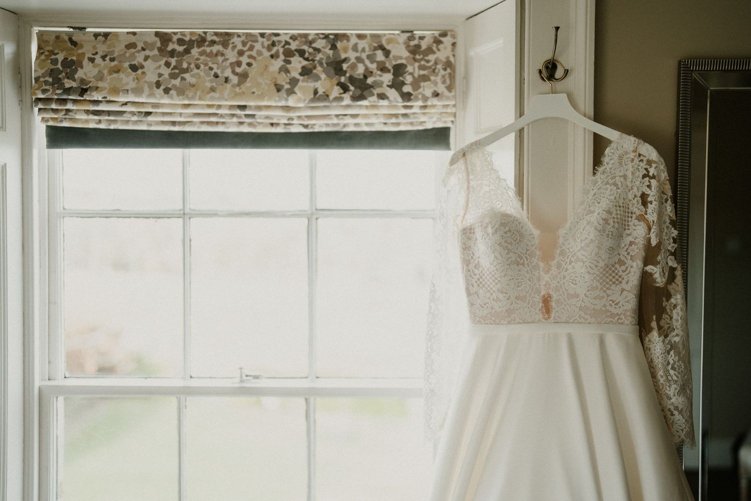 Wedding dress hanging by window with natural light streaming through white curtains.