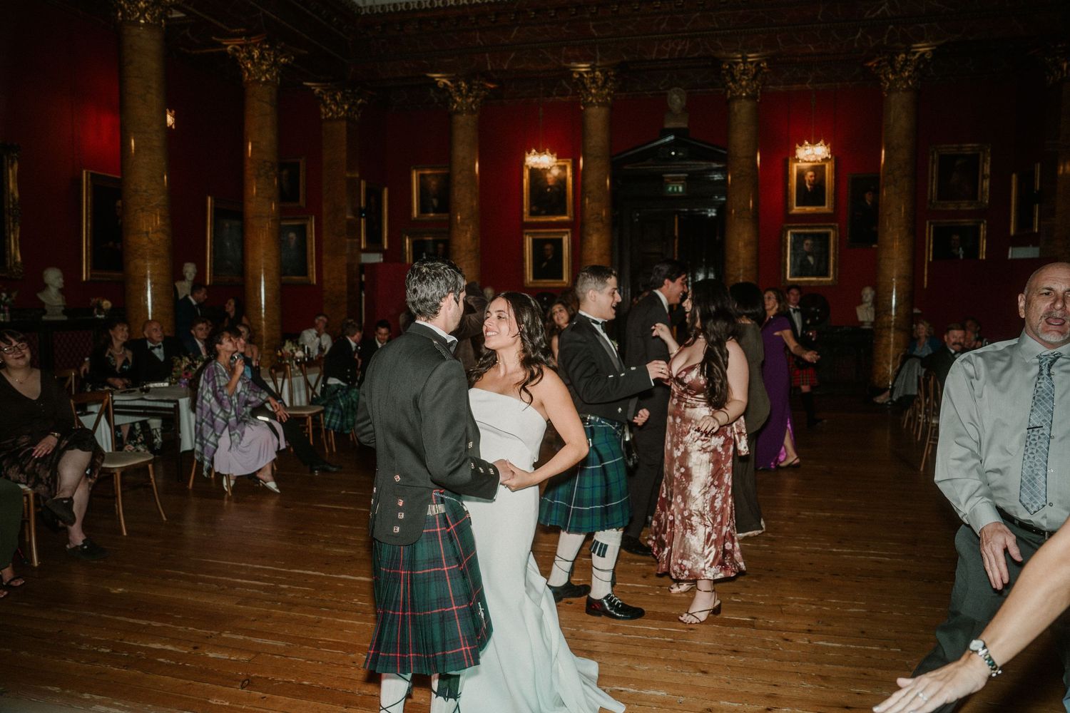 Wedding celebration with dancing guests in a historic venue featuring red walls and classical architecture.