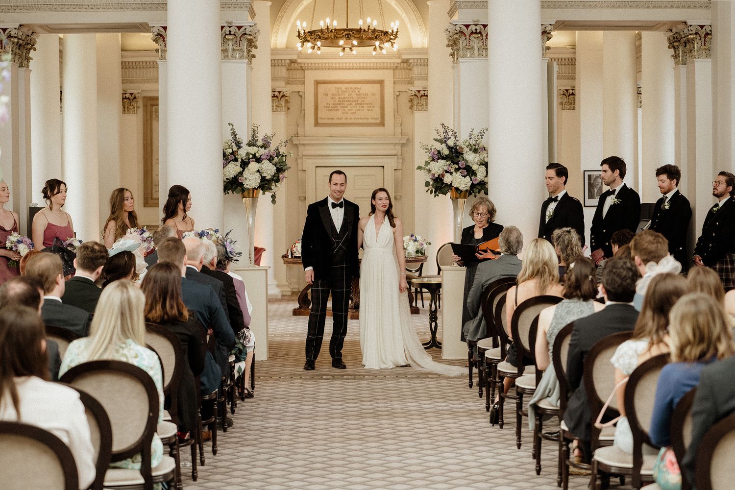 Wedding ceremony taking place in an elegant ballroom with white decor and chandelier lighting.
