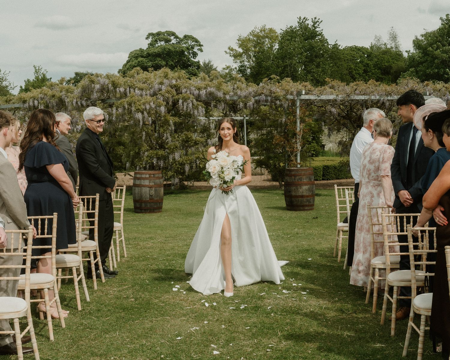 Bride walks down outdoor aisle in elegant gown at garden wedding ceremony with wisteria backdrop.