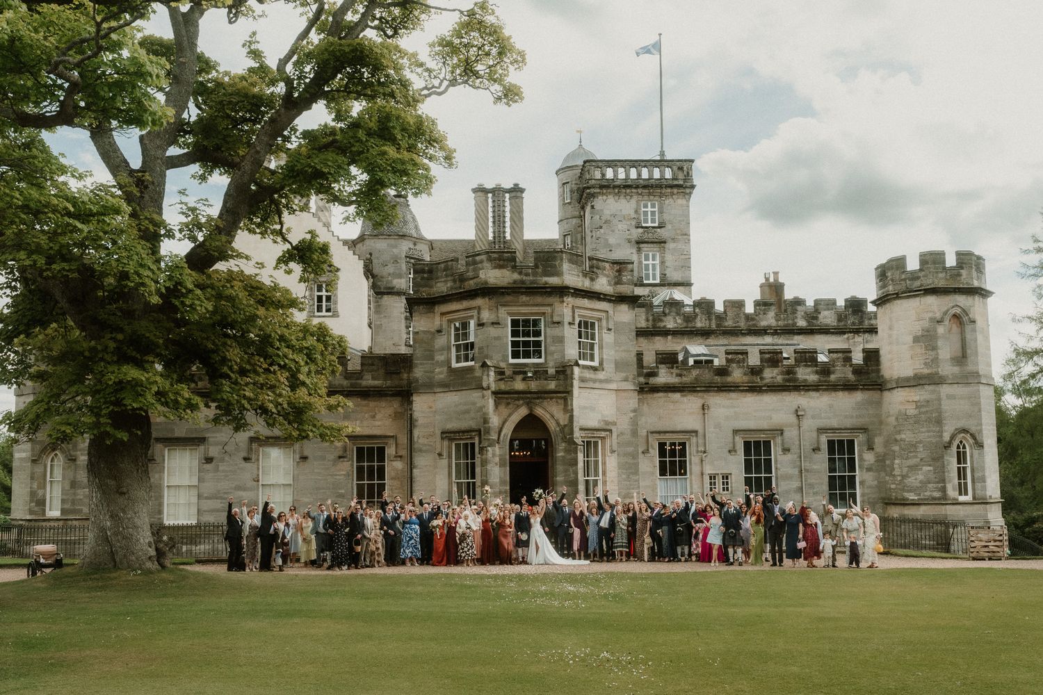 Wedding party posing in front of a grand Scottish castle venue with lush green grounds and cloudy sky.
