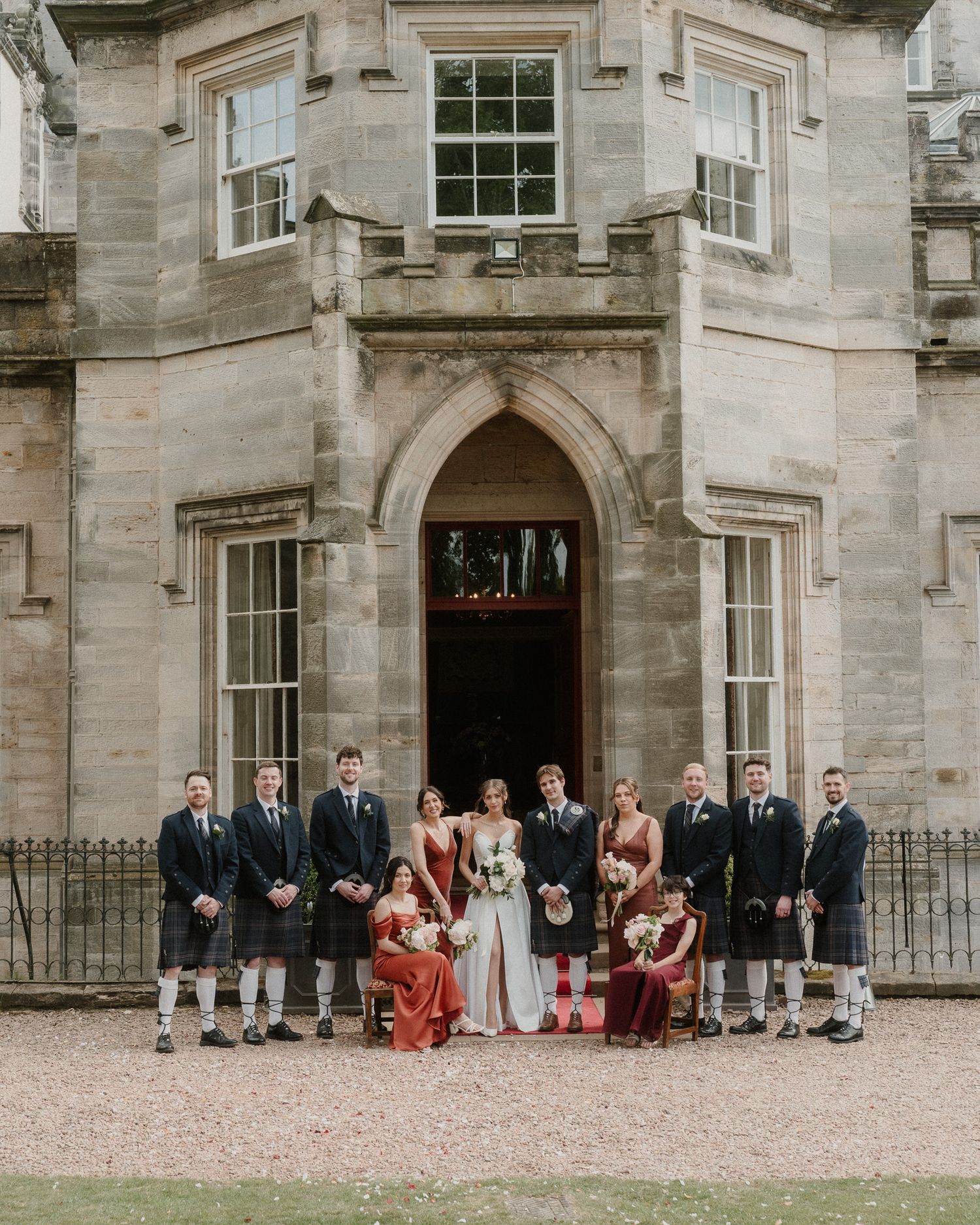 Wedding party poses before a grand Scottish stone manor, groomsmen in kilts and bridesmaids in red gowns.