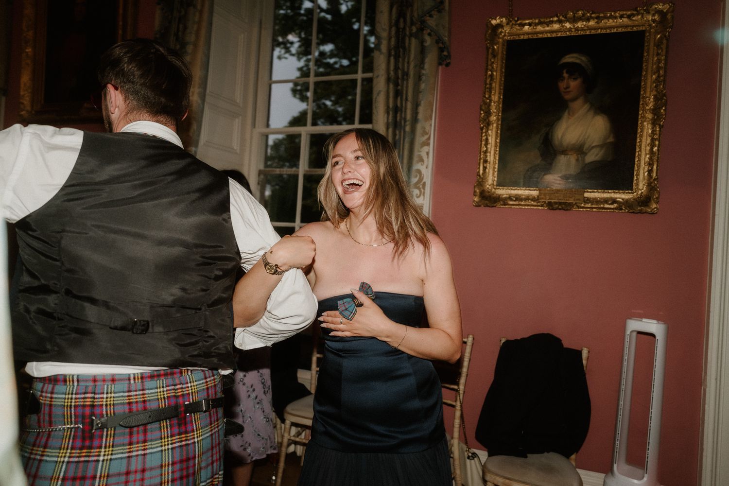 Guests dancing joyfully at a Scottish wedding reception, woman in navy dress laughing beside man in tartan kilt.