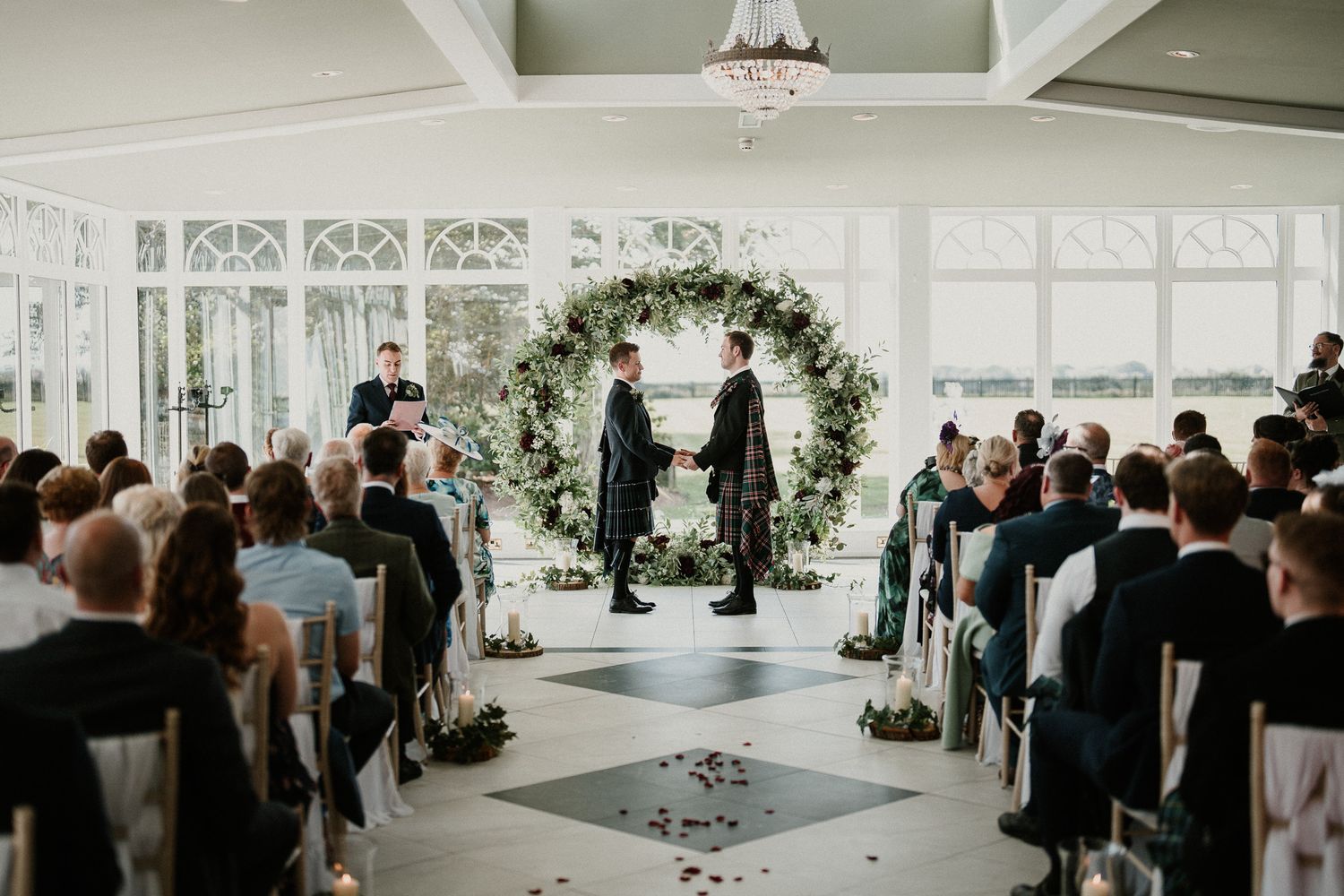 A wedding ceremony in a bright indoor venue with a floral arch and seated guests in formal attire.