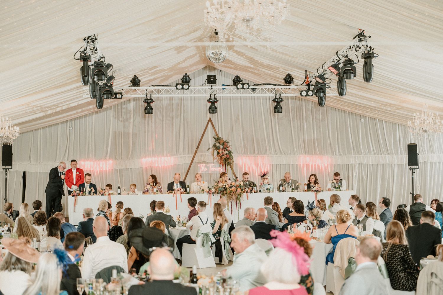 A large wedding reception in a white draped tent with hanging floral arrangements and chandeliers.
