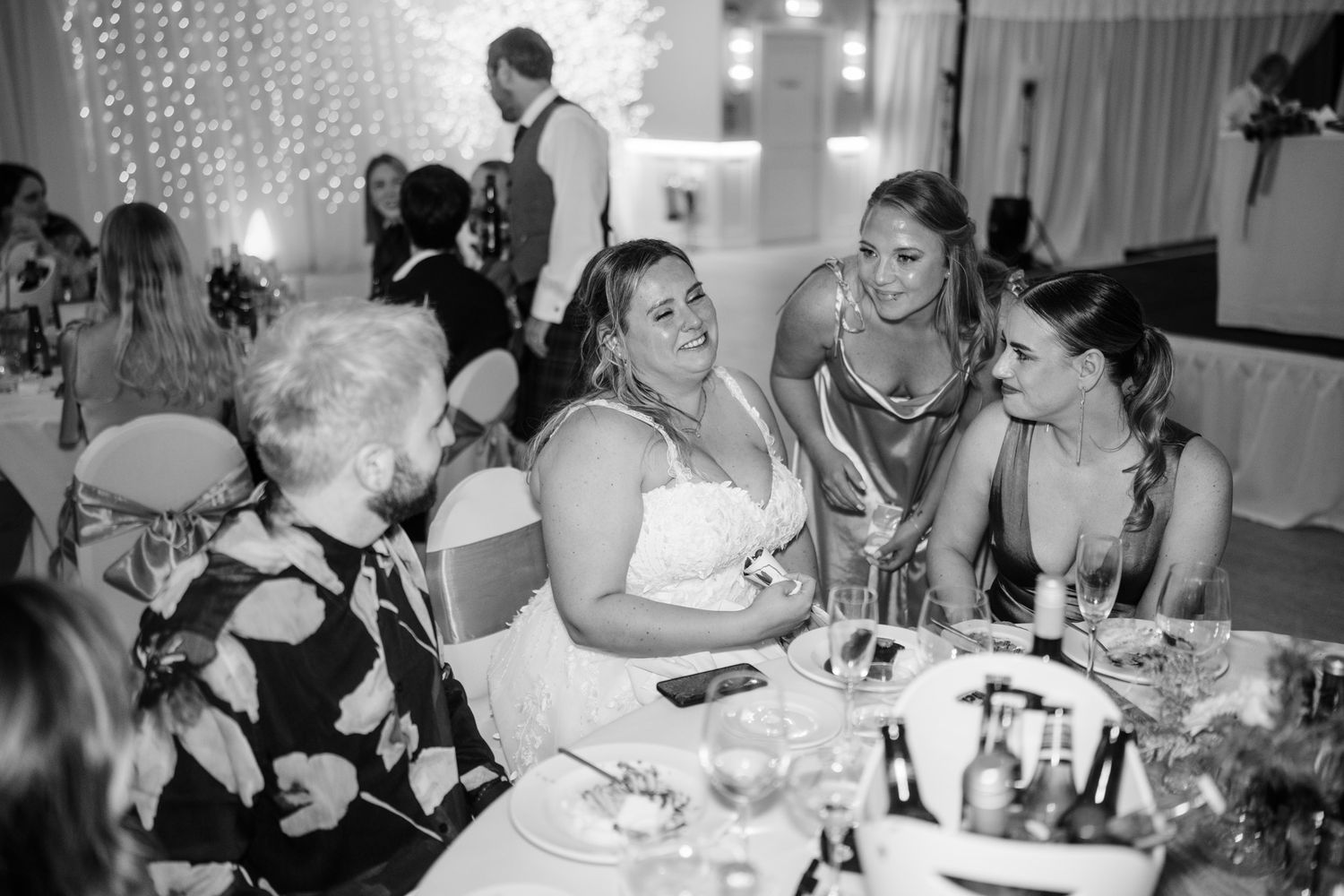 Black and white photo of wedding reception guests sharing conversation at decorated table.