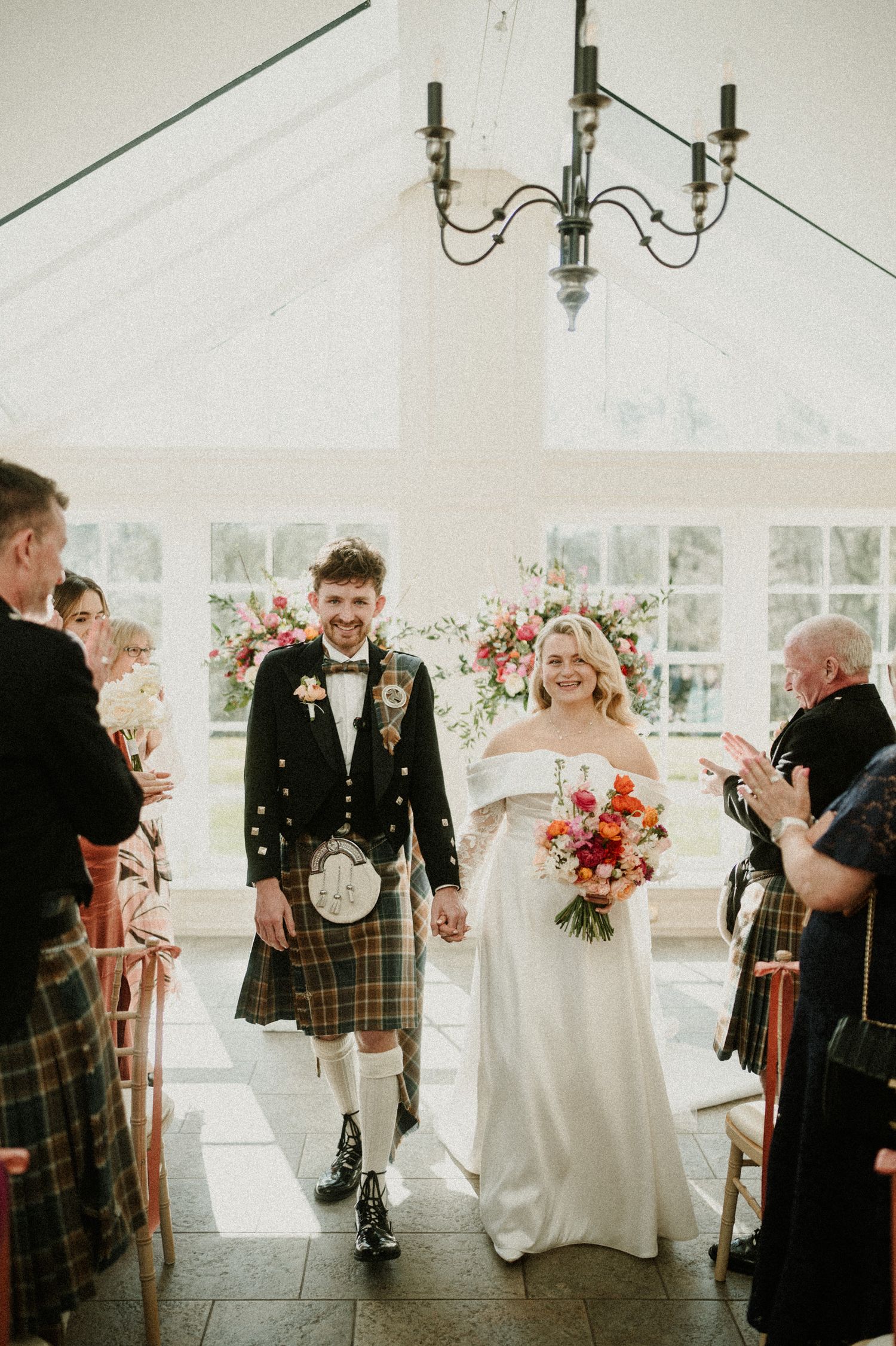 Newlyweds walk down the aisle, groom in traditional Scottish kilt and bride in elegant white gown.